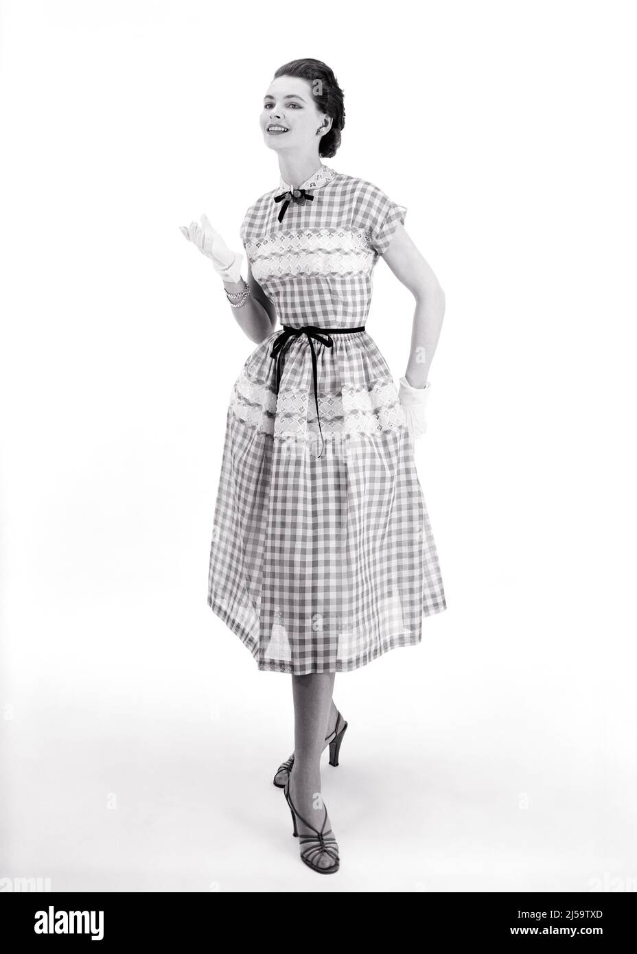 1950S BRÜNETTE MODELL TRÄGT KARIERTES KURZARM-KLEID MIT SPITZEN-BANNER AUF ROCK UND BÜSTE BAND KRAWATTE AN TAILLE UND HALS - F3873 CRR001 HARS BÜSTE UND KARIERTE BERUFE POSIEREN STILVOLLE MODELLIERUNG MODE MODELLIERUNG POSE TAILLE JUNGE ERWACHSENE FRAU BANNER SCHWARZ UND WEISS ALTMODISCH Stockfoto