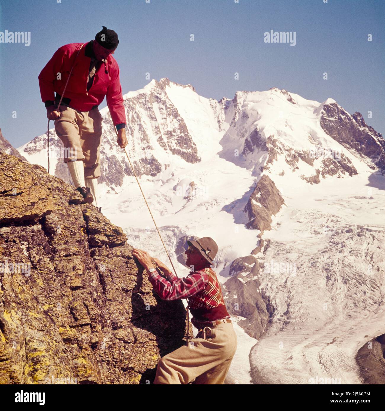 1960S ZWEI MÄNNER KLETTERN IN DEN SCHWEIZER ALPEN MIT SEILEN ZUM AUFSTIEG VON FELSVORSPRÜNGEN SCHNEEBEDECKTE BERGE UND GLETSCHER IM HINTERGRUND - KC3065 LUT001 HARS VORSPRUNG UND AUFSTIEG MIT SEILEN BEDECKTE GLETSCHER ALPEN FLEXIBILITÄT MUSKELN HELFEN BERGSTEIGERN SCHWEIZER KAUKASISCHE ETHNIE ALTMODISCH Stockfoto