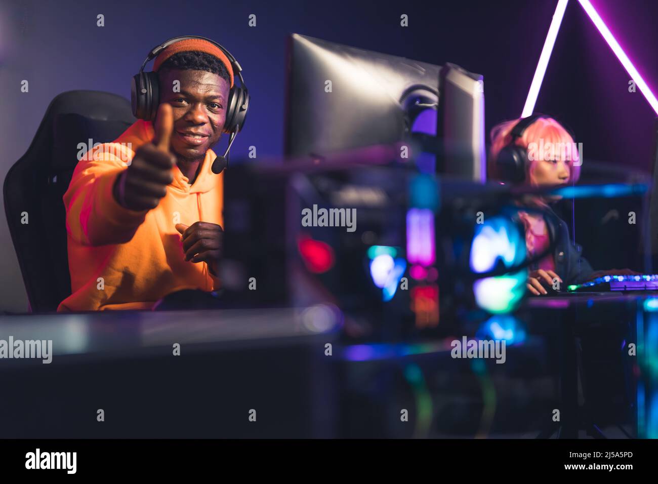 Fröhlicher Afro-Amerikaner in einem orangenen Hoodie, der in einem Cyber-Café sitzt, ein weiteres Spiel mit seinen Gefährten gewinnt und den Daumen hoch zur Kamera zeigt. Hochwertige Fotos Stockfoto
