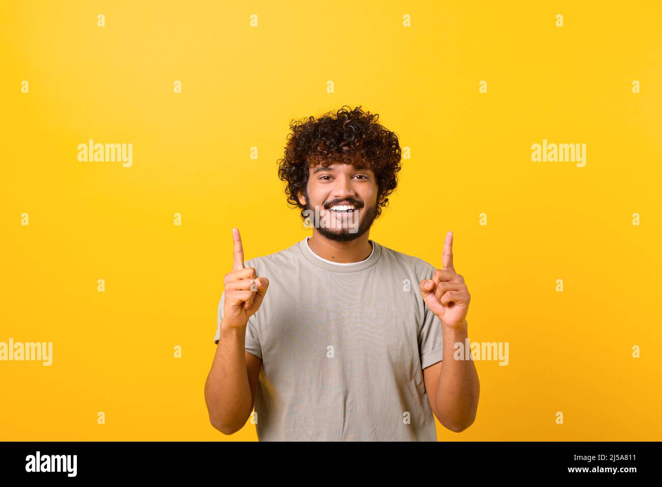 Attraktiver indischer Mann mit glücklicher Gesichtsausdruck, der mit beiden Fingern nach oben zeigt und den Kopierraum präsentiert. Innenaufnahme des Studios isoliert auf gelbem Hintergrund Stockfoto