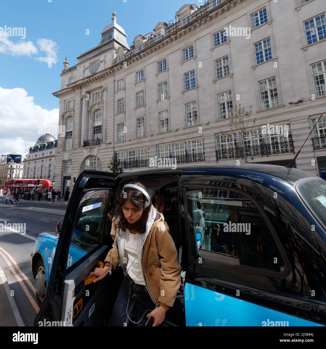 London, Greater London, England, April 09 2022: Kunde mit Kopfhörern, der mit einem Bus im Hintergrund aus einem Taxi oder Taxi in der Regent Street aussteigen muss. Stockfoto