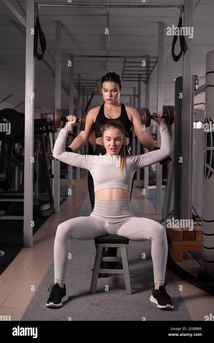 Aufnahme einer muskulösen jungen Frau, die Hanteln im Fitnessstudio hebt und mit einem persönlichen Trainer trainiert. Die Dame pumpt ihre Schultermuskeln mit einem Himmel auf Stockfoto