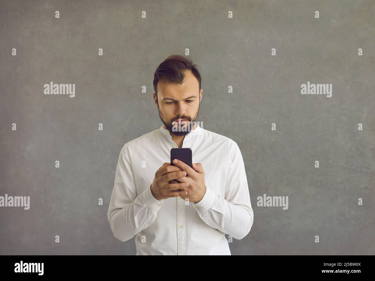 Ruhiger bärtiger kaukasischer Geschäftsmann mit Smartphone, der vor grauem Hintergrund steht. Stockfoto