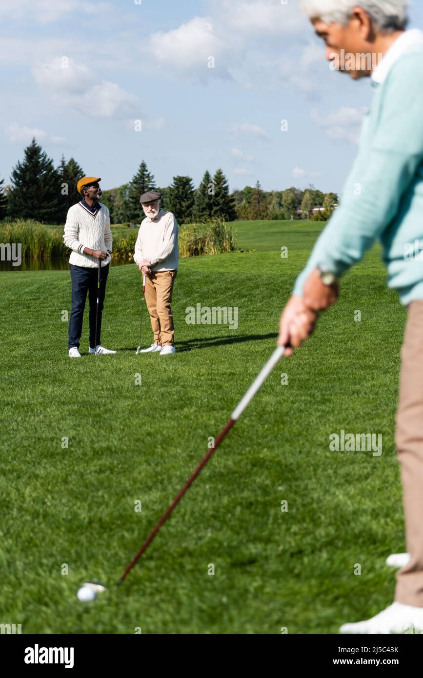 Ältere interracial Männer in flachen Kappen Blick auf asiatische Freund Golf spielen auf verschwommenem Vordergrund Stockfoto