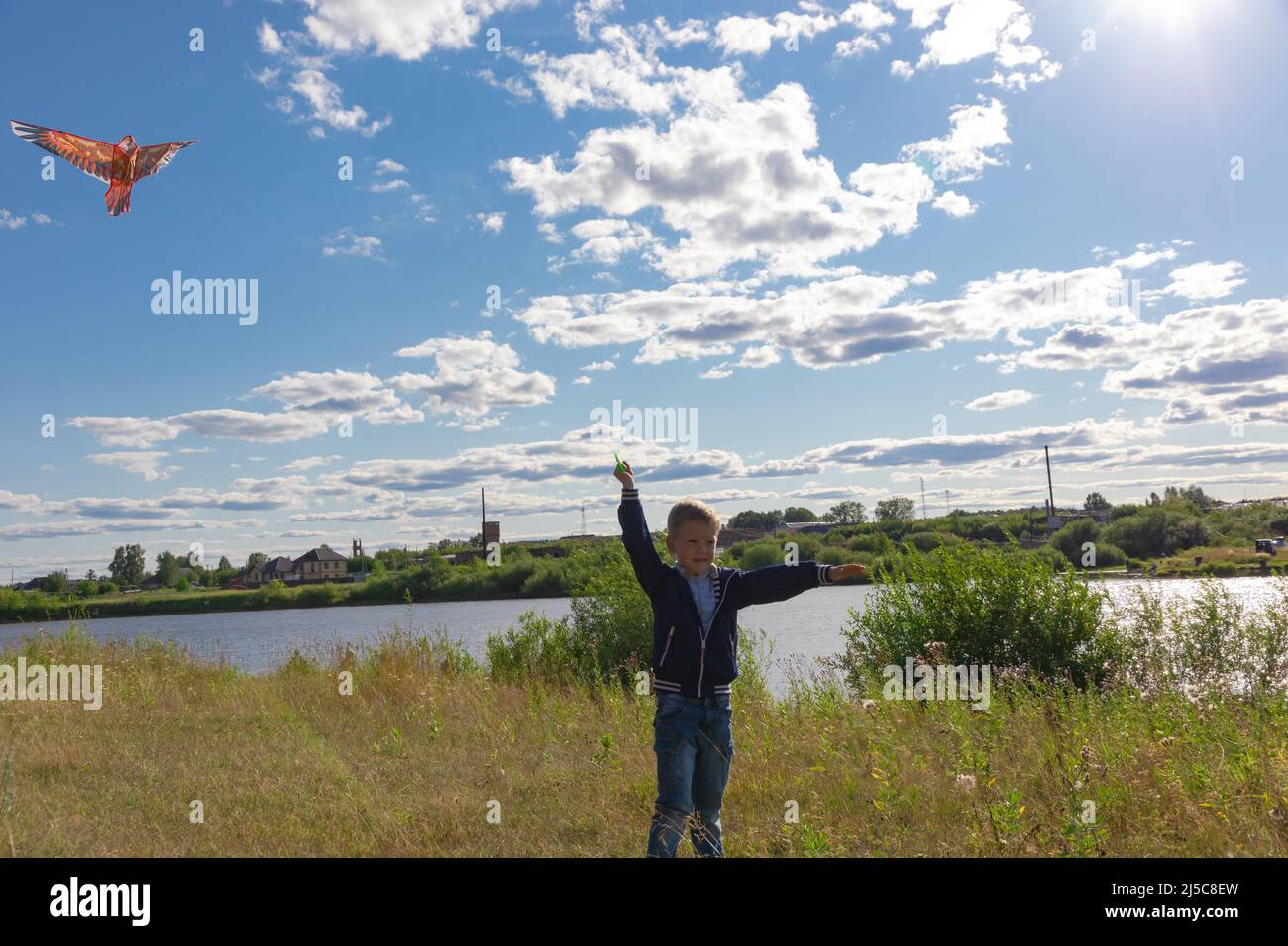 Ein sechsjähriger Vorschuljunge in blauer Jacke lässt an einem Sommertag vor dem Hintergrund eines klaren blauen Himmels einen Drachenvögel in die Natur fliegen. Die helle Stockfoto