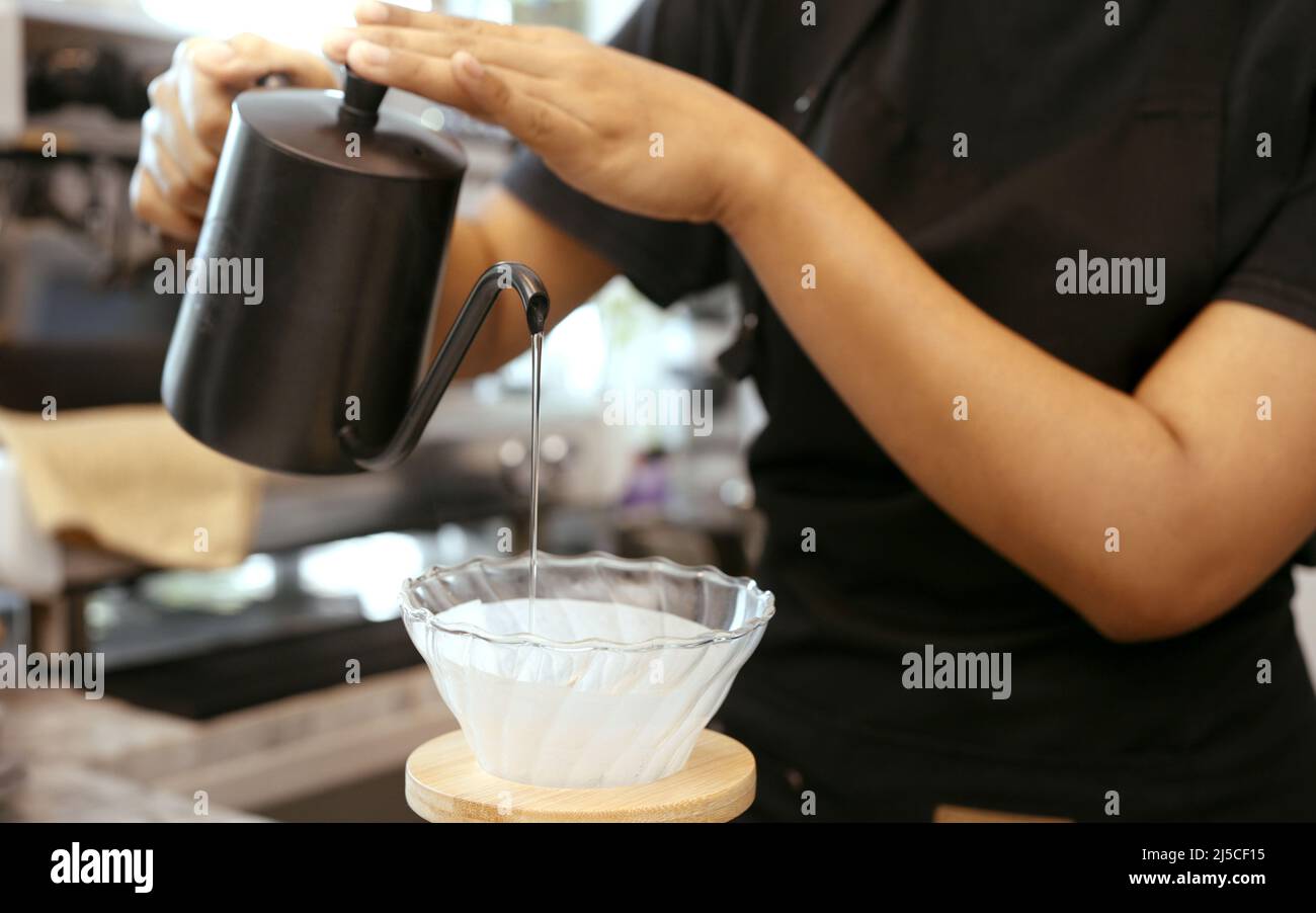 Eine Cafébetreiberin, die eine Schürze trägt, gießt heißes Wasser über den gerösteten Kaffeesatz, um Kaffee für die Kunden im Laden zuzubereiten. Stockfoto