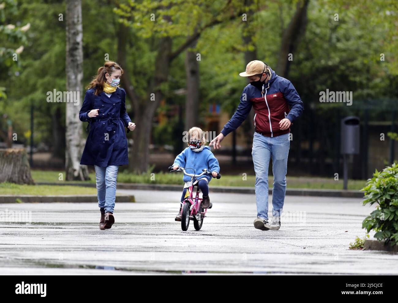 Endlich raus - Familie trägt Gesichtsmasken in einem öffentlichen Park, während sie mit ihrer kleinen Tochter Fahrrad fahren. [Automatisierte Übersetzung] Stockfoto