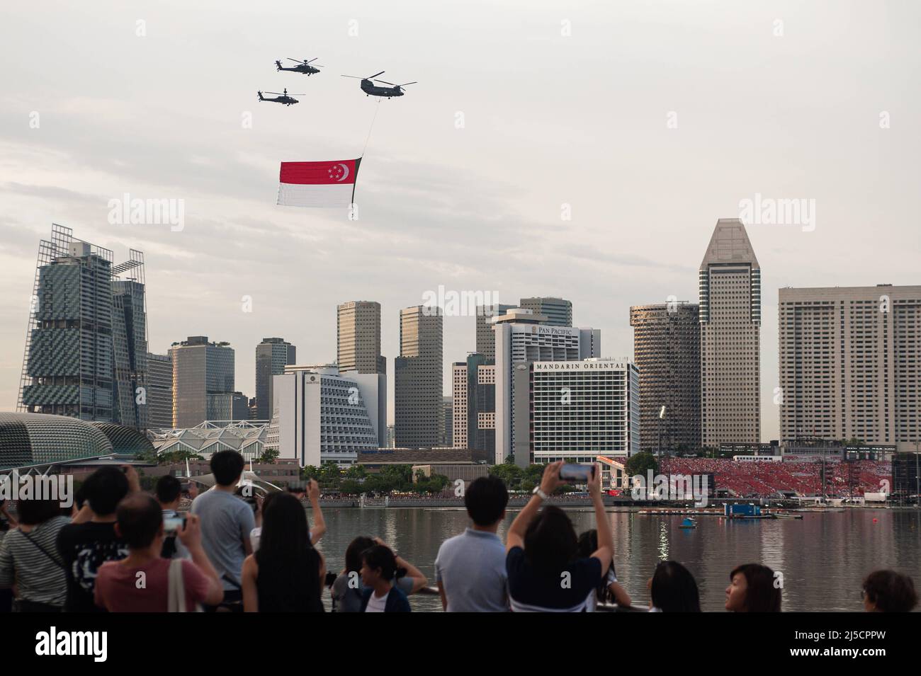 07/21/2018, Singapur, Republik Singapur, Asien - Zuschauer beobachten, wie Hubschrauber der Luftwaffe (RSAF) während eines Trainingsfluges eine riesige Staatsflagge über Wolkenkratzer in Marina Bay fliegen, um sich auf den bevorstehenden Nationalfeiertag am 9. August vorzubereiten. [Automatisierte Übersetzung] Stockfoto