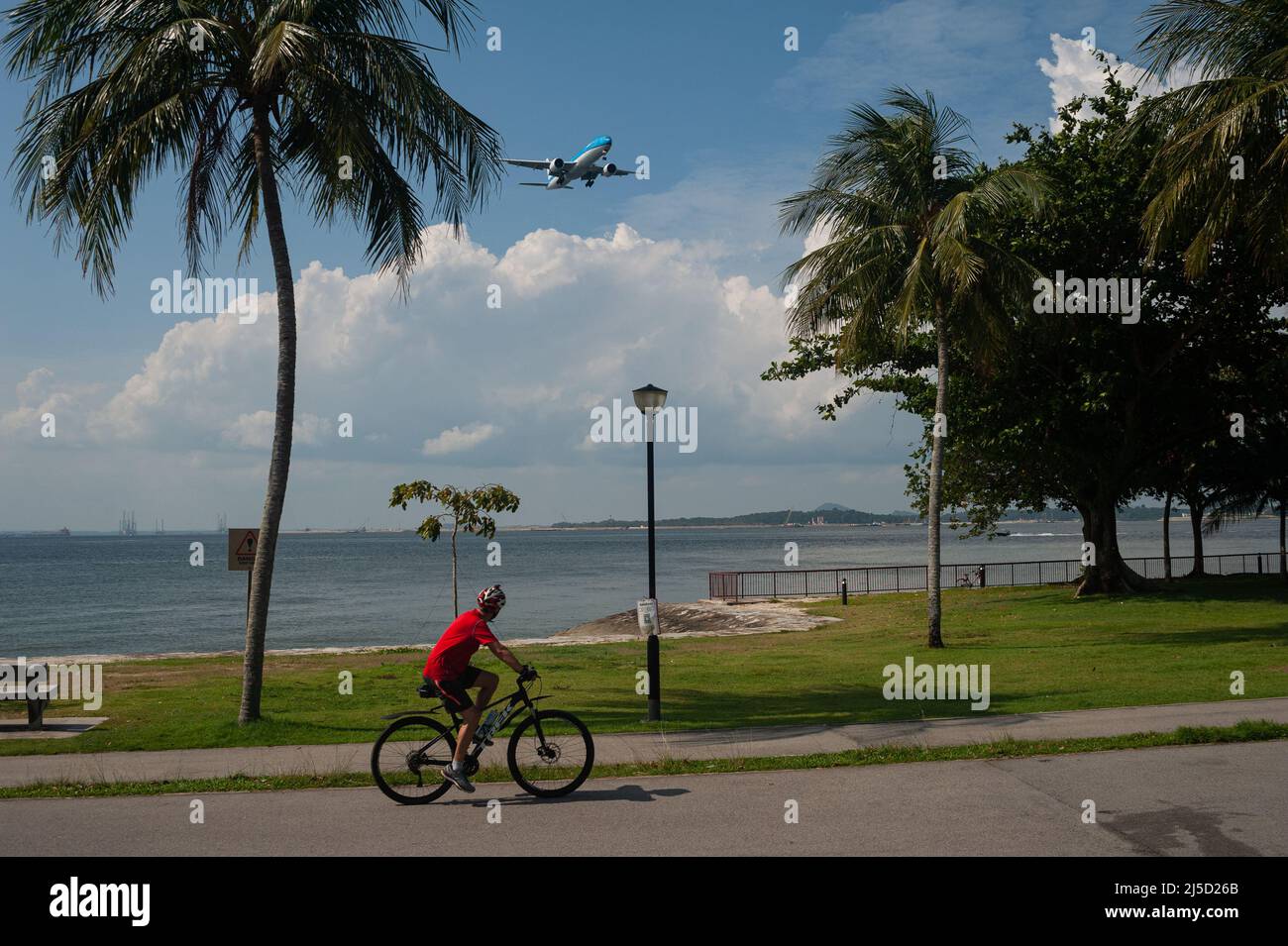 07/08/2021, Singapur, Republik Singapur, Asien - Ein Mann fährt während der anhaltenden Corona-Krise mit dem Fahrrad am Ufer des Changi Beach Parks entlang, während ein Flugzeug im Hintergrund auf dem Weg zum Changi International Airport ist. [Automatisierte Übersetzung] Stockfoto