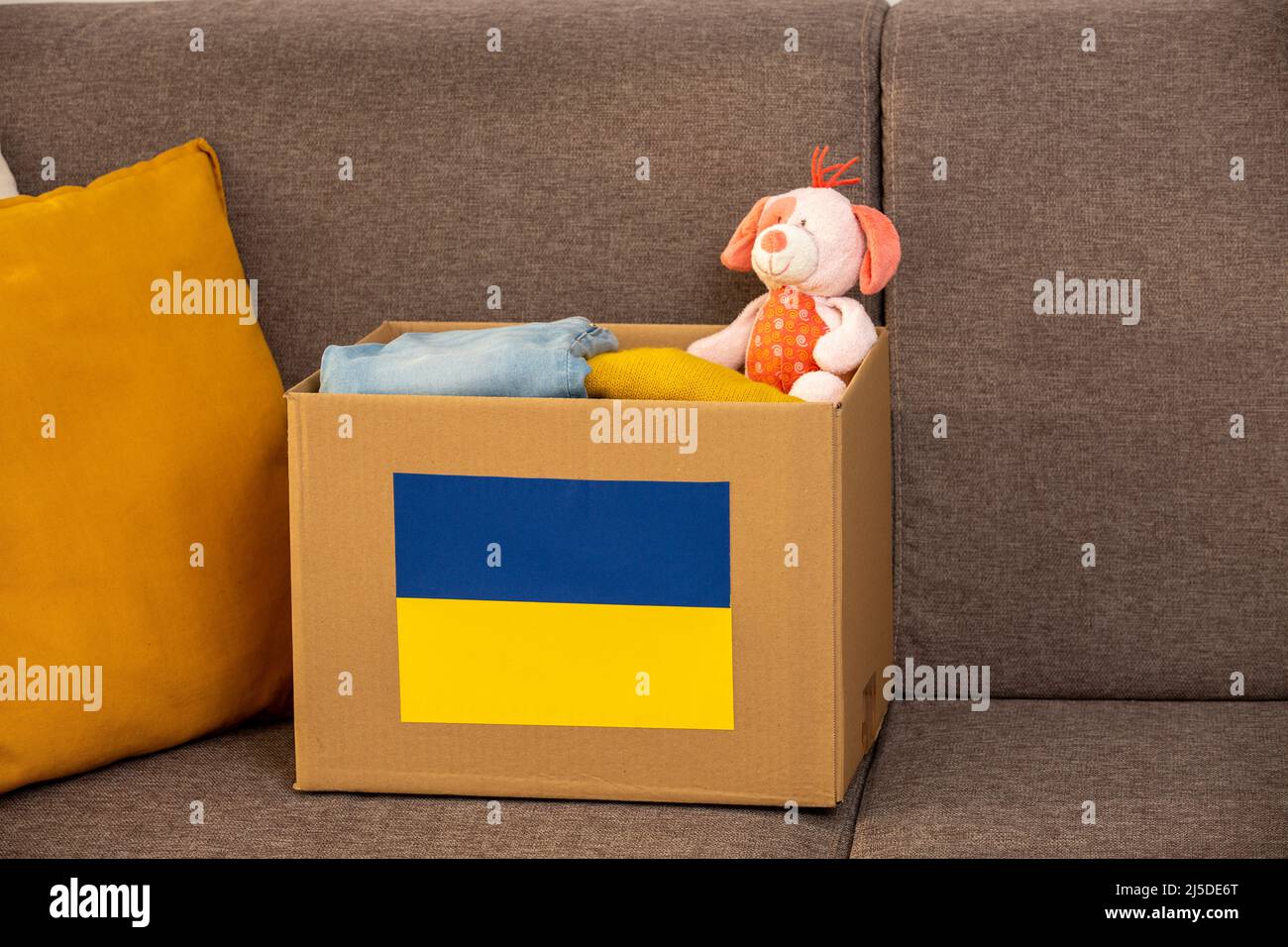 Nahaufnahme-Box mit ukrainischer Flagge für humanitäre Hilfe Spende mit Kindersachen und Spielzeug zu Hause auf dem Sofa.Bleiben Sie in der Ukraine. Humanitäre Hilfe für Stockfoto