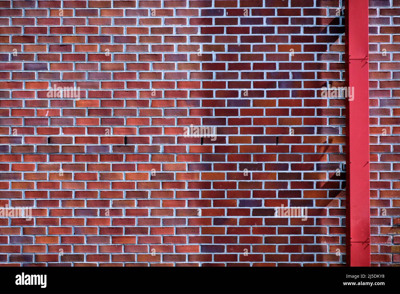 Ziegelwand mit Ziegel in roten Farbtönen von orange bis lila. Die rote Dachrinne kreuzt den Ziegel und sorgt so für einen Kontrast. Stockfoto