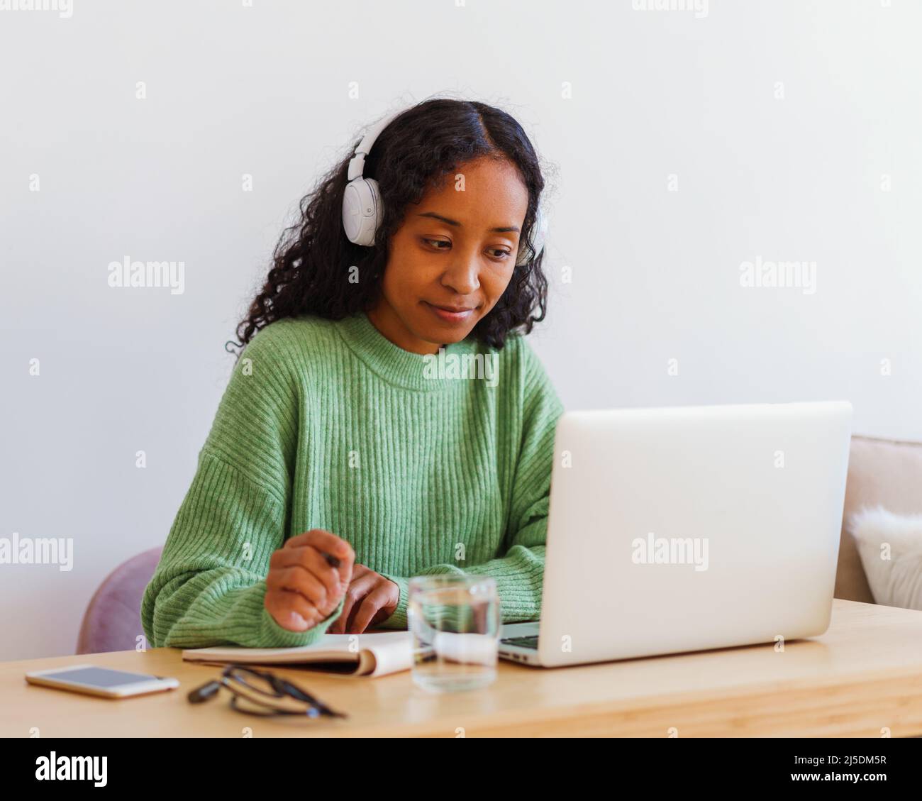 Junge afroamerikanische Frau, die mit Kopfhörern an einem Laptop arbeitet, während sie sich Notizen im Notizbuch notiert Stockfoto