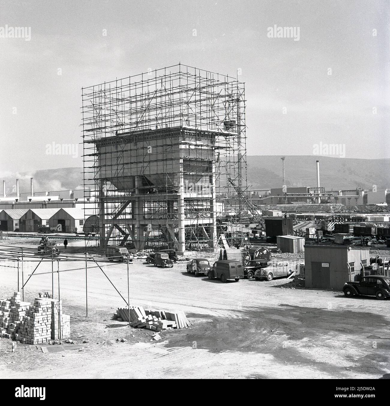 1950, historische Bauarbeiten beim Bau eines Betonwasserturms im riesigen Stahlwerk Abbey der Steel Company of Wales in Port Talbot, Wales, Großbritannien. Ein Schild auf der korragierten Hütte auf dem Boden in ihrer Nähe besagt: 'Temporäre Umspannstation'. Autos und Lieferwagen der damaligen Zeit stehen auf dem Parkplatz. Erbaut auf dem Gelände des alten Margam Eisen- und Stahlwerks, wurde das Abteiwerk 1951 eröffnet. Bis zum Jahr 1953, bis zum Jahr 1960s, war es das größte Stahlwerk in Europa. 1967 wurde das Unternehmen verstaatlicht und wurde Teil von British Steel. Stockfoto