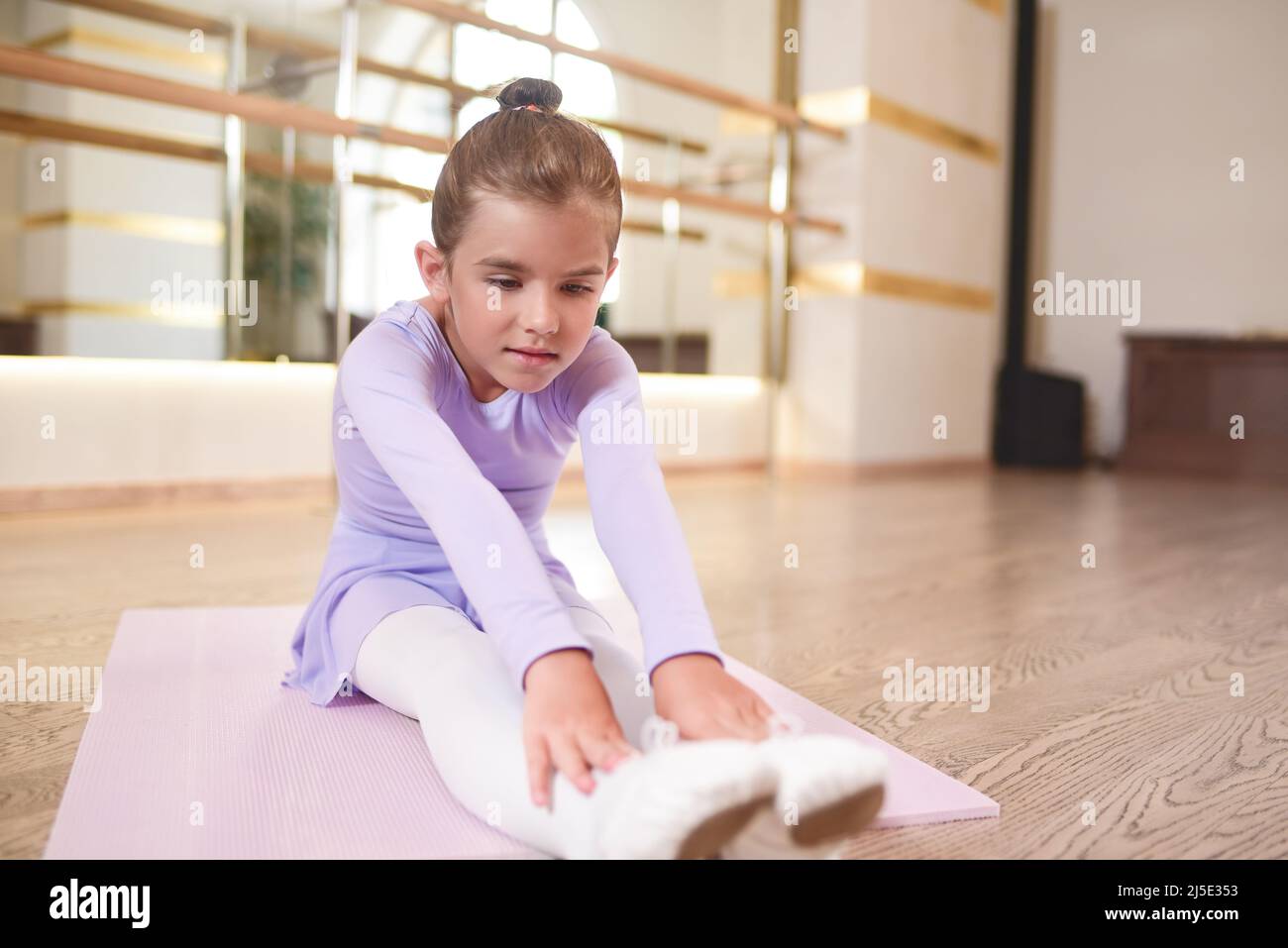 Entzückendes Foto der kleinen Ballerina in einem lila Kleid und weißen Strumpfhosen Stretching-Übungen in einem Saal mit großen Spiegeln. Stockfoto