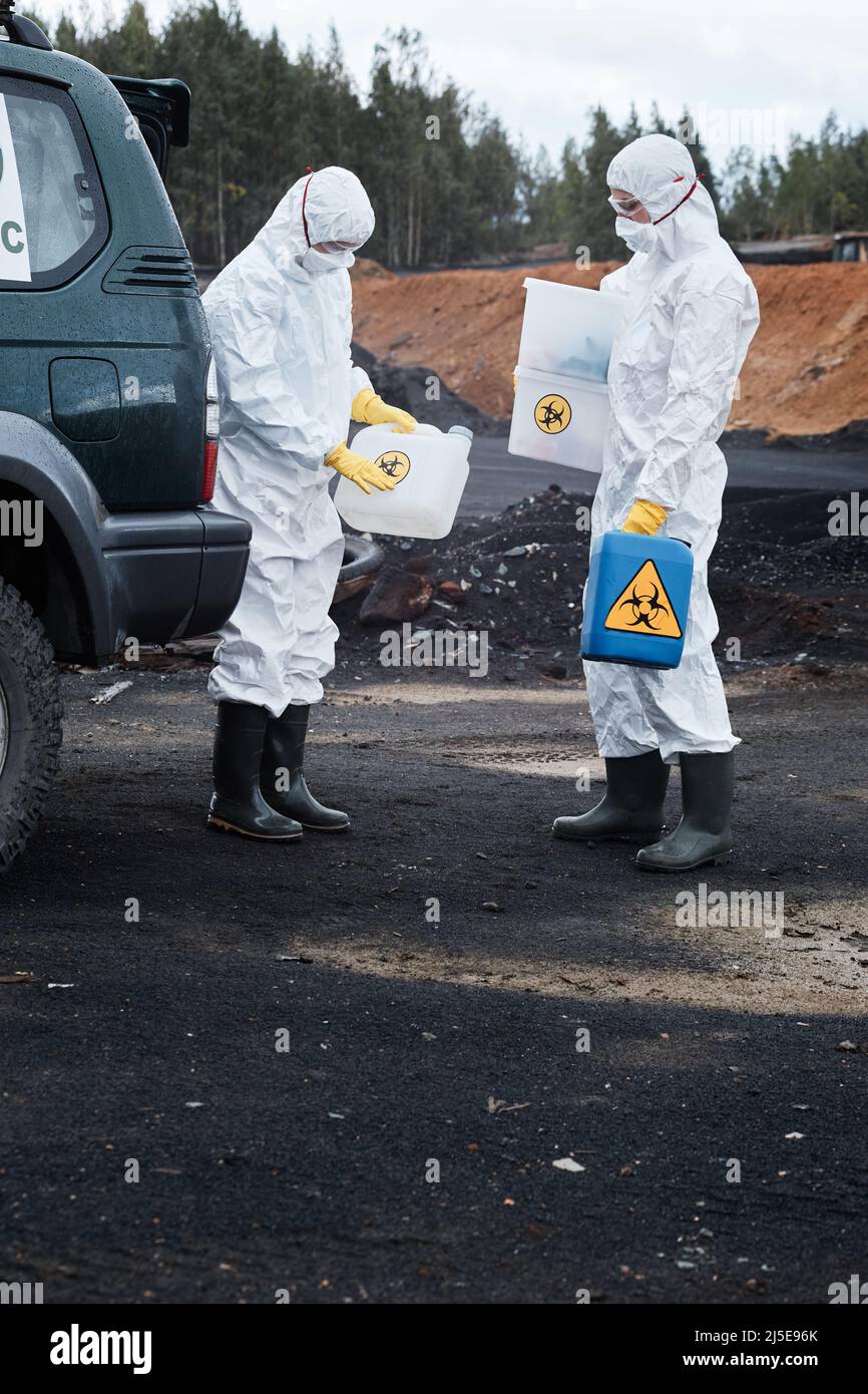 Zwei Ökologen in Arbeitskleidung und Gummistiefeln, die auf verschmutztem Land am Auto stehen und Container für giftige Substanzen halten Stockfoto