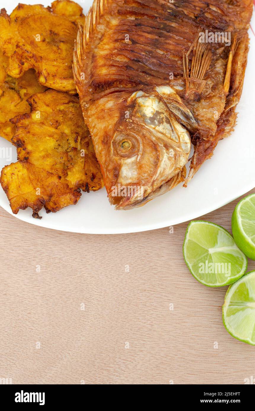 Gebratener Mojarra-Fisch, knusprig, traditionell im kolumbianischen Pazifik. Begleitet von Patakonen mit Haus und Zitrone, auf einem hellen Holzhintergrund.Copy Space. Stockfoto