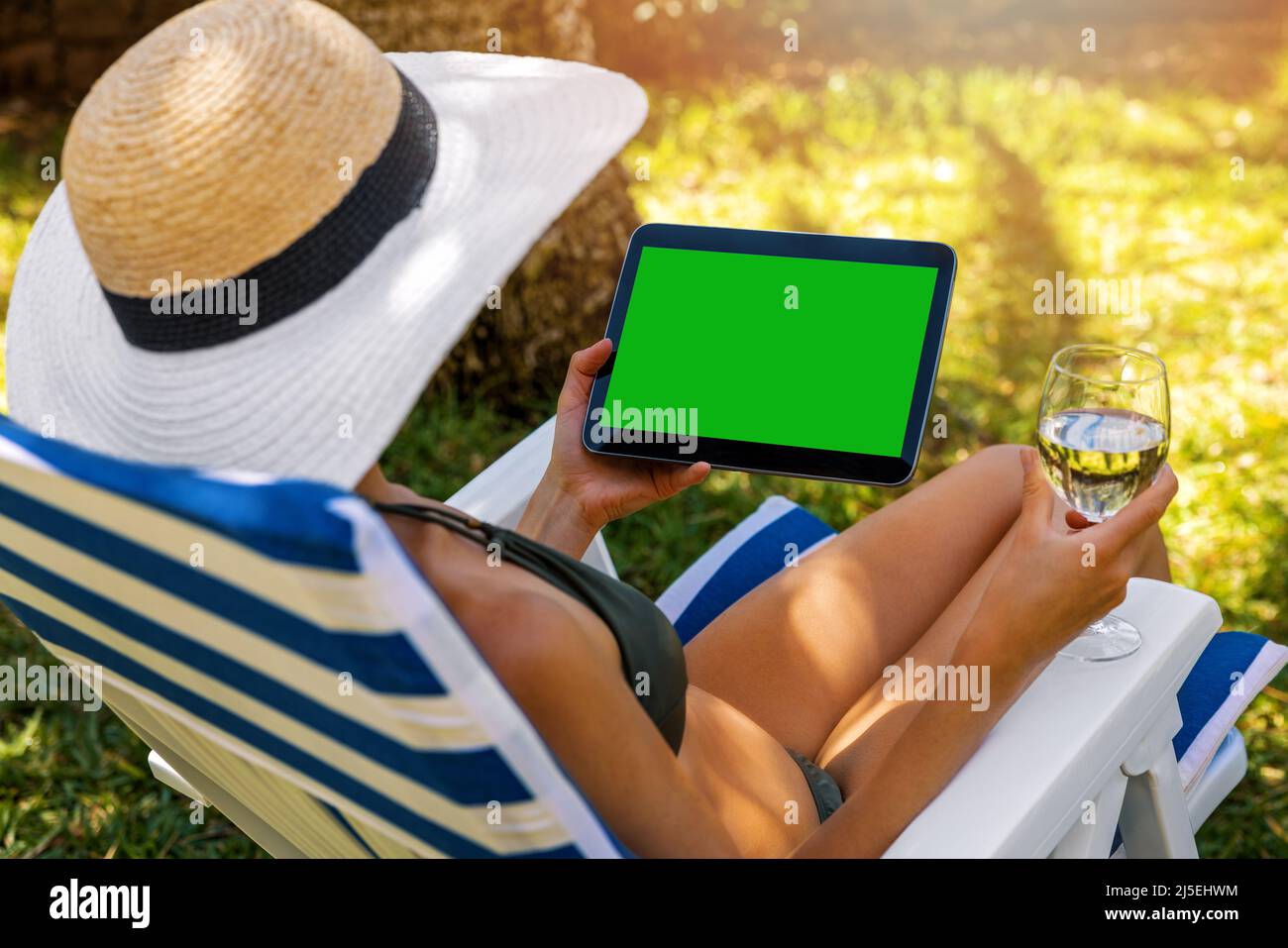 Nachbau einer Frau im Bikini mit digitalem Tablet und Weintrinken, während sie sich in der Sonnenliege im Garten entspannt Stockfoto