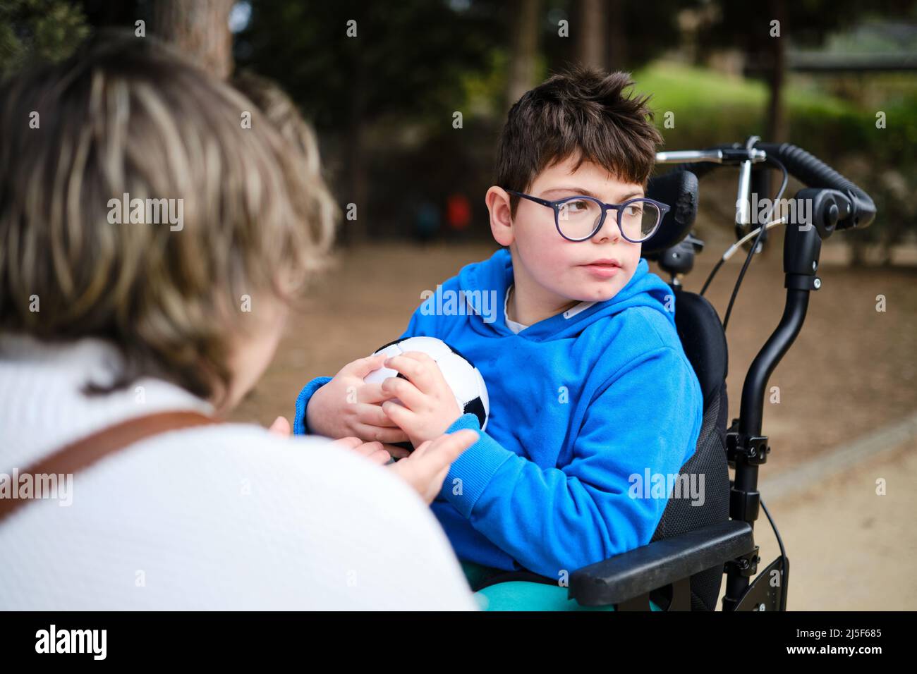 Behinderter Junge im Rollstuhl mit einem Fußball, während er mit seiner Mutter im Park spielt. Stockfoto