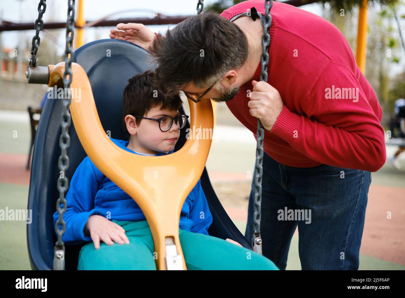 Ein behinderter Junge und sein Vater spielen gerne auf dem Spielplatz des Parks mit einer Schaukel für besondere Bedürfnisse. Stockfoto