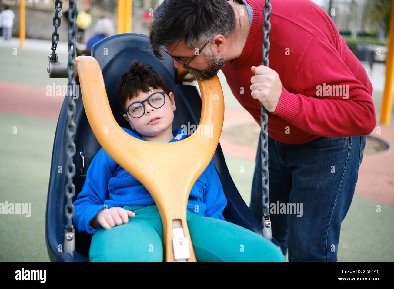 Behinderter Junge und sein Vater genießen es gemeinsam, während das Kind auf einer speziellen Schaukel im Park schaukelt. Stockfoto