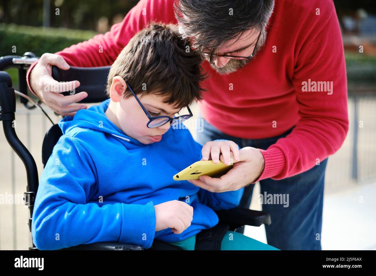 Behindertes Kind im Rollstuhl und sein Vater, der ein Mobiltelefon während eines Spaziergangs im Freien verwendet. Stockfoto