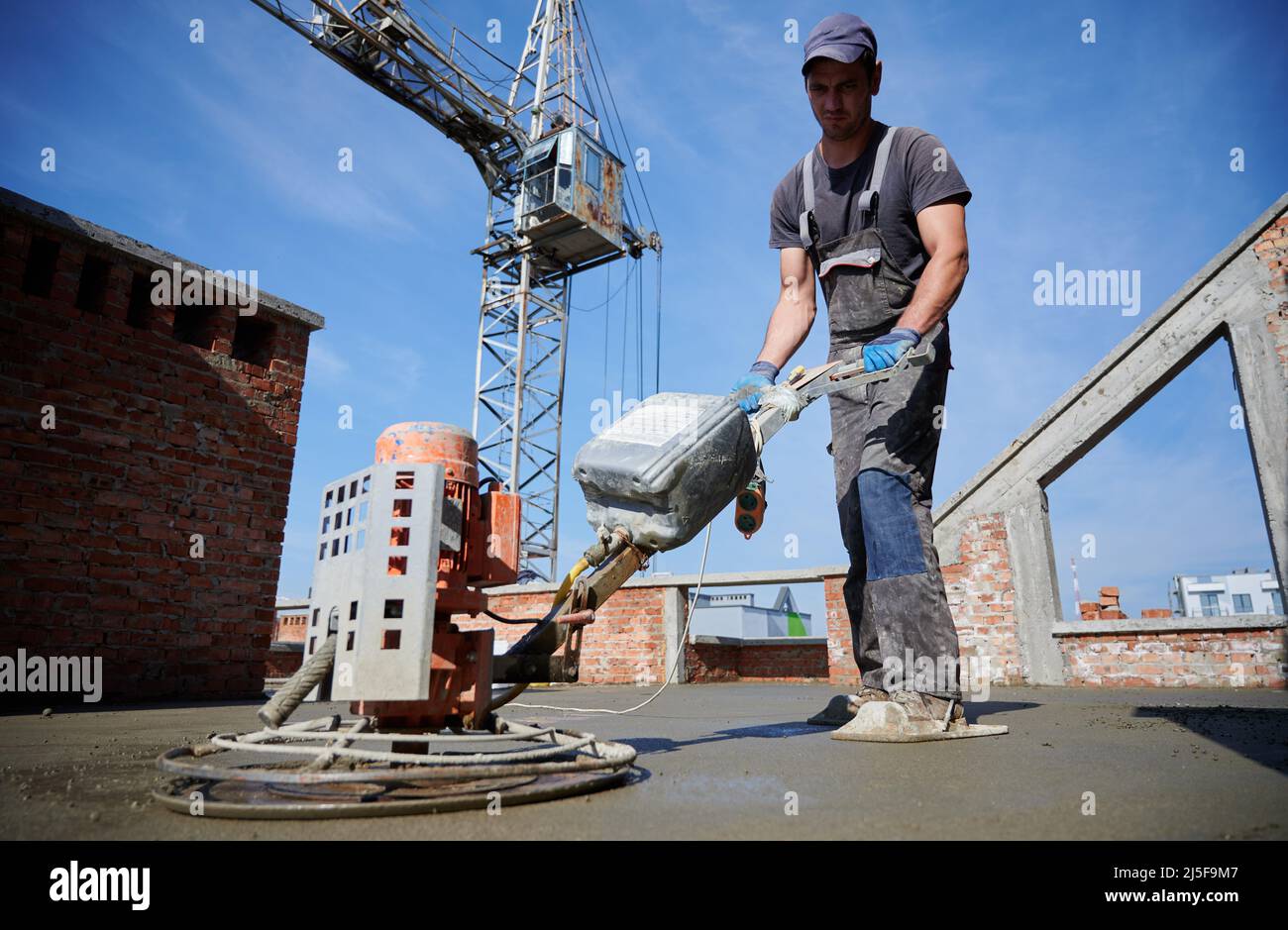 Spezialist in der Anordnung des Bodens, der die Ungleichheit der Oberfläche nach der Installation des halbtrockenen Estrich von der speziellen Maschine nivelliert. Auf dem Hintergrund Turmdrehkran und blauer wolkenloser Himmel. Stockfoto