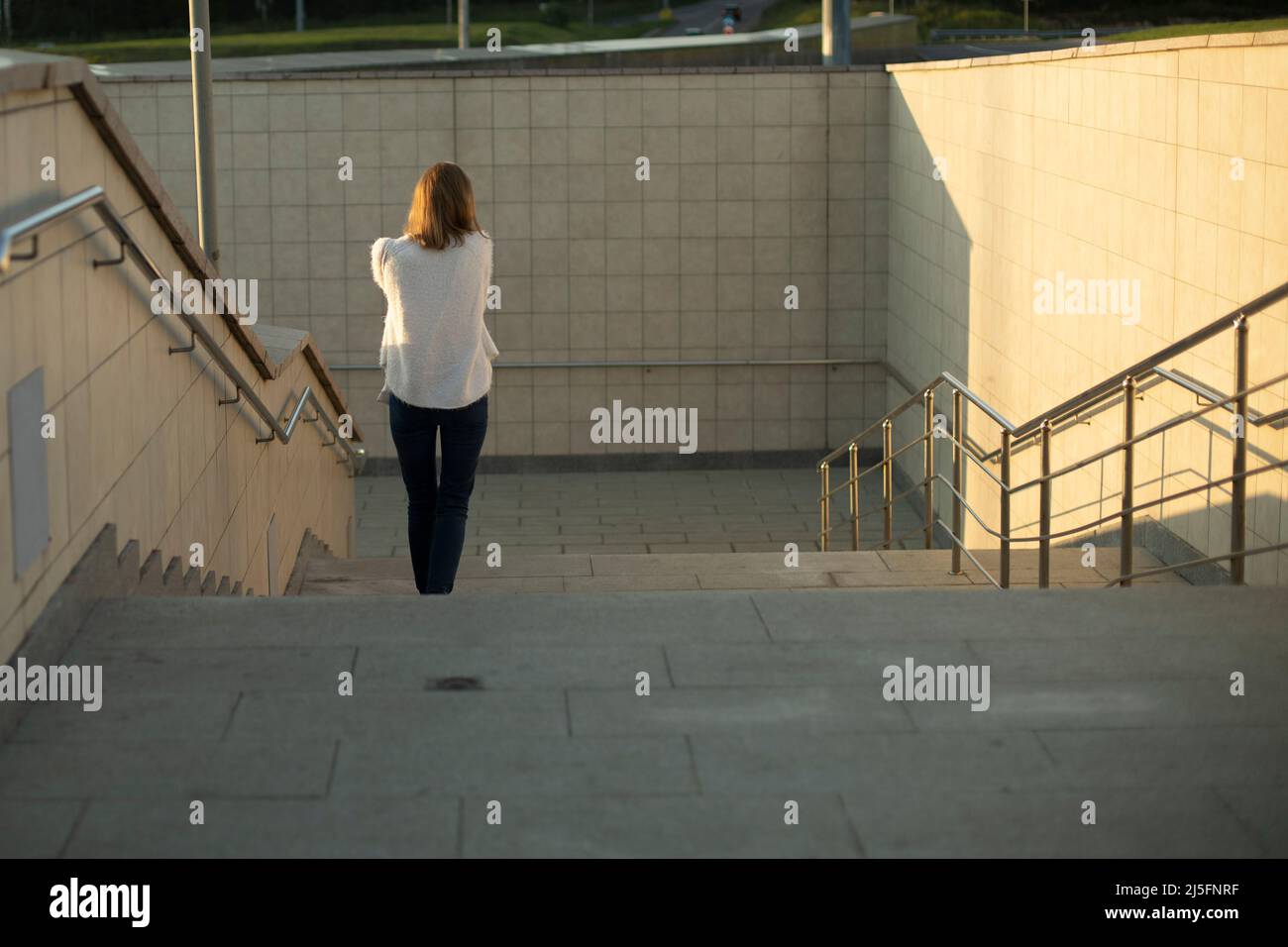 Mädchen stolpert die Treppe hinunter. Mädchen steigt in eine unterirdische Fußgängerüberführung über den Highway. Frau im weißen Pullover geht die Stufen hinunter. Untergehende Sonne shi Stockfoto