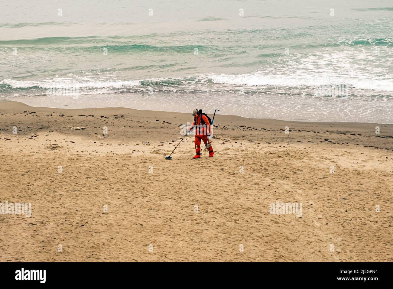 Ein Mann mit einem Metalldetektor geht in einem roten Anzug am Strand entlang. Zeitlupe, 4K, Zeitlupe Stockfoto