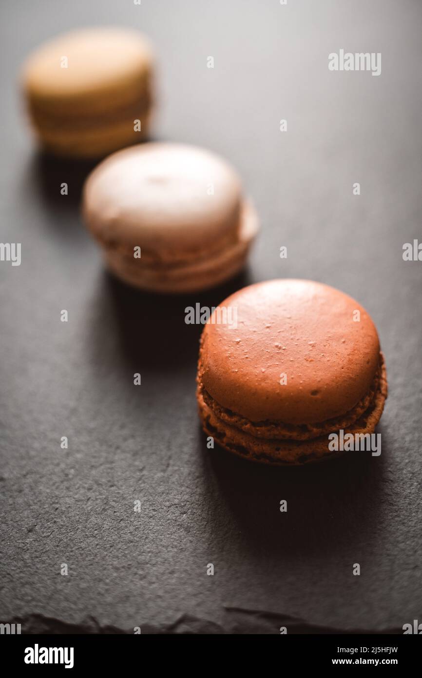 Ein dunkles, launiges Bild von Schokolade, Karamell und Vanillemakronen, die auf einer Schiefertafel auf einem dunkelgrauen, strukturierten Betonhintergrund angeordnet sind. Stockfoto