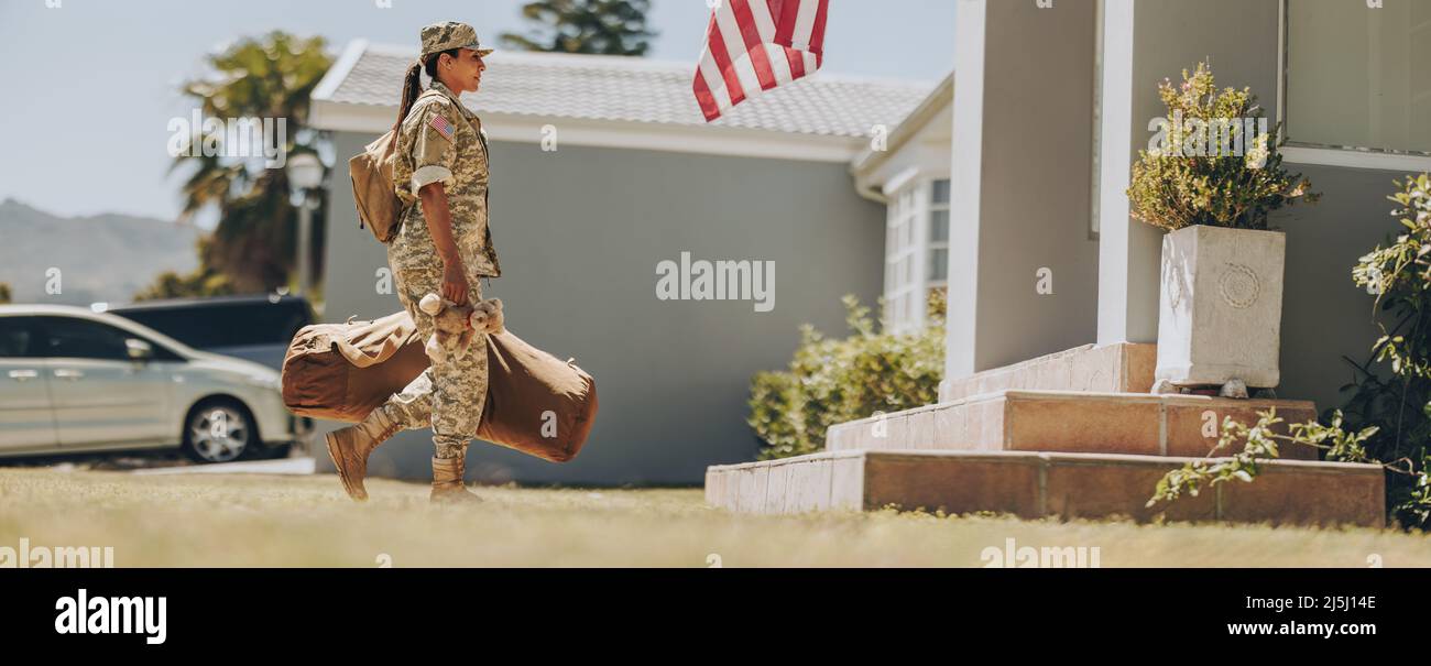 Mutige Militärmom, die einen Teddybär in der Hand hält, während sie mit ihrem Gepäck zu ihrem Haus geht. Amerikanische Soldatin, die nach dem Dienst h nach Hause zurückkehrt Stockfoto