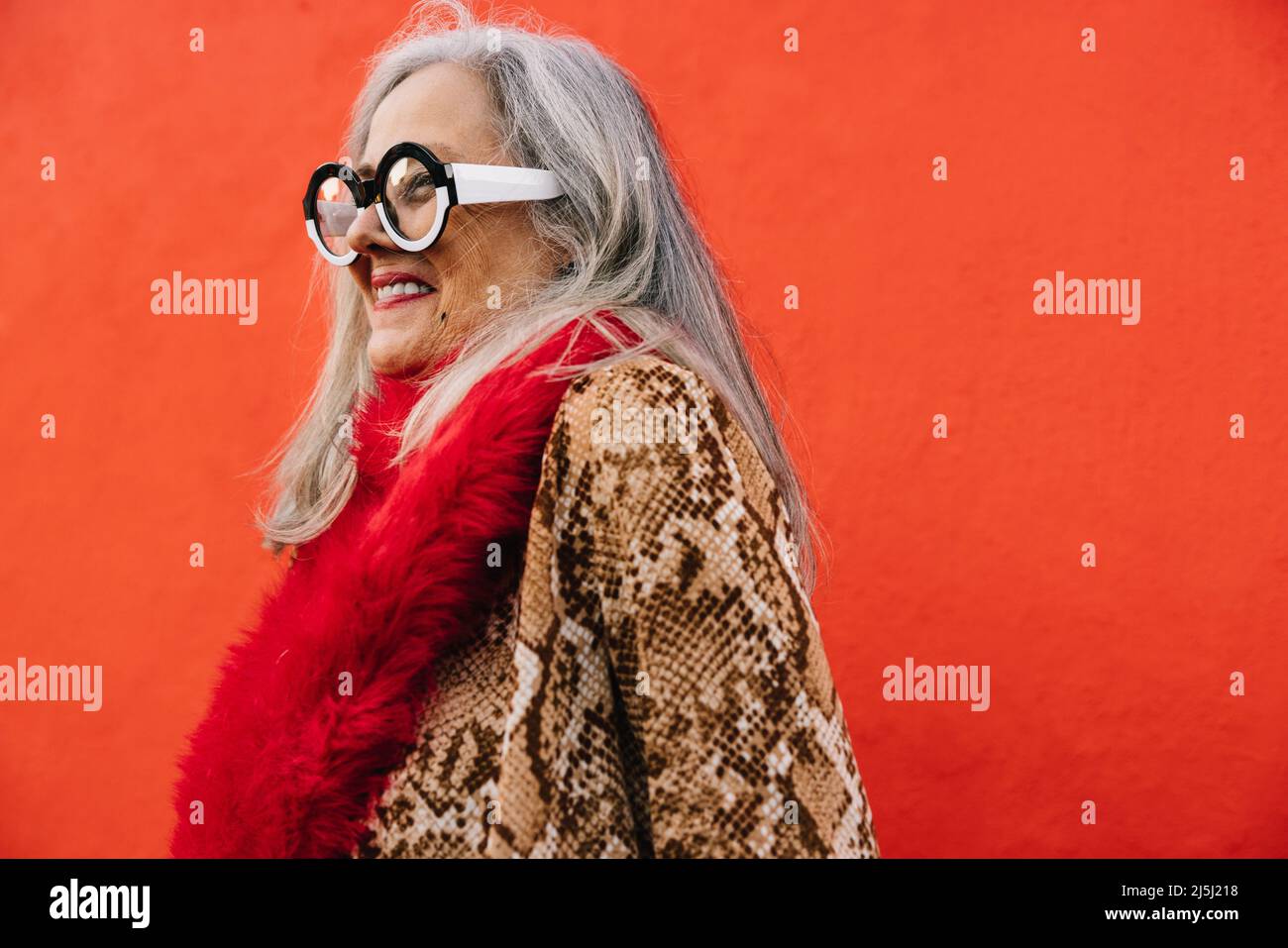Fröhliche ältere Frau, die fröhlich lächelt, während sie vor einem roten Hintergrund steht. Sorglose ältere Frau, die sich in farbenfrohen Kleidungen jugendlich und lebendig fühlt Stockfoto
