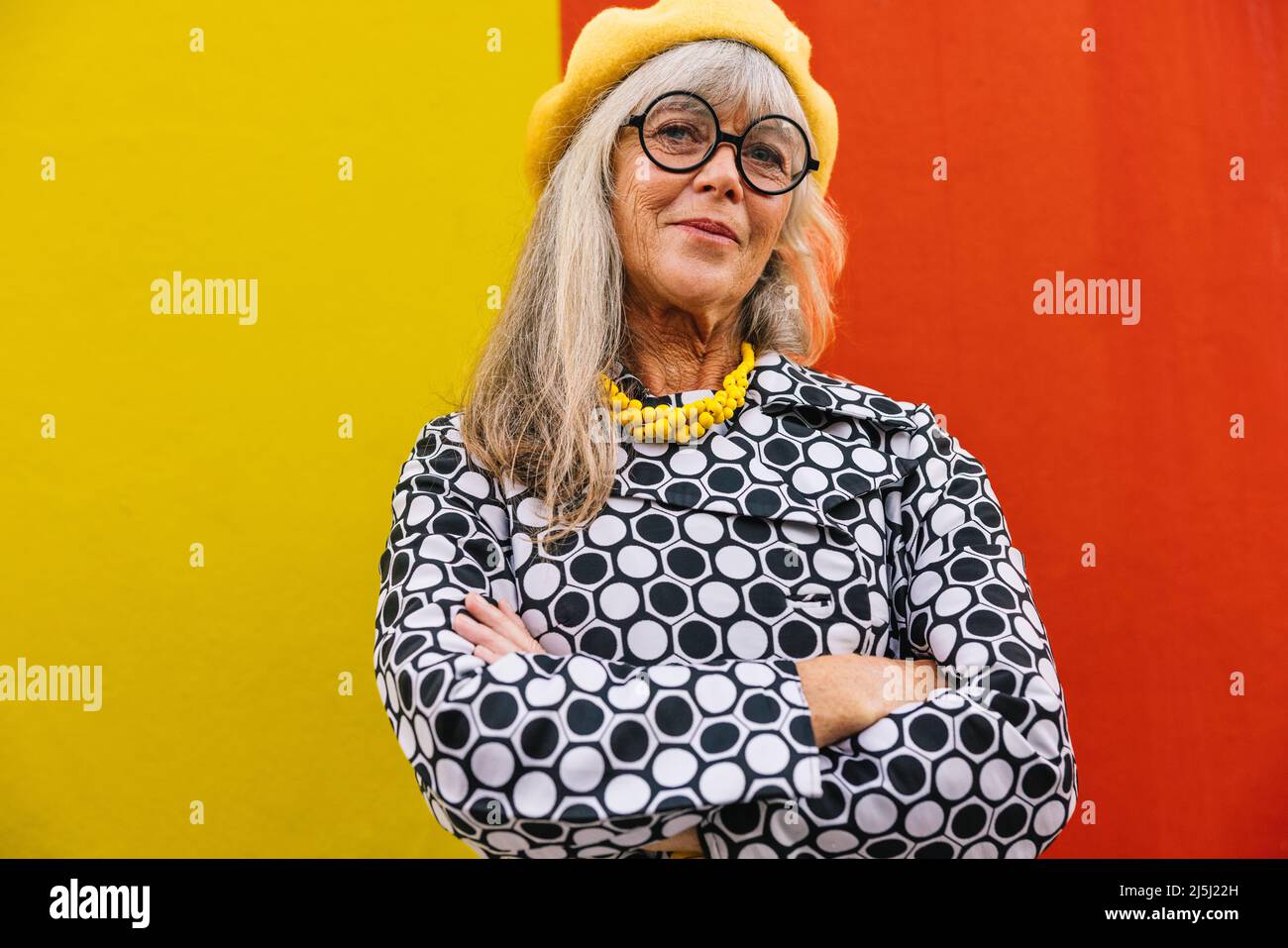 Selbstbewusste, reife Frau, die mit gekreuzten Armen auf die Kamera schaut. Stilvolle ältere Frau trägt bunte Kleidung gegen eine rote und Stockfoto