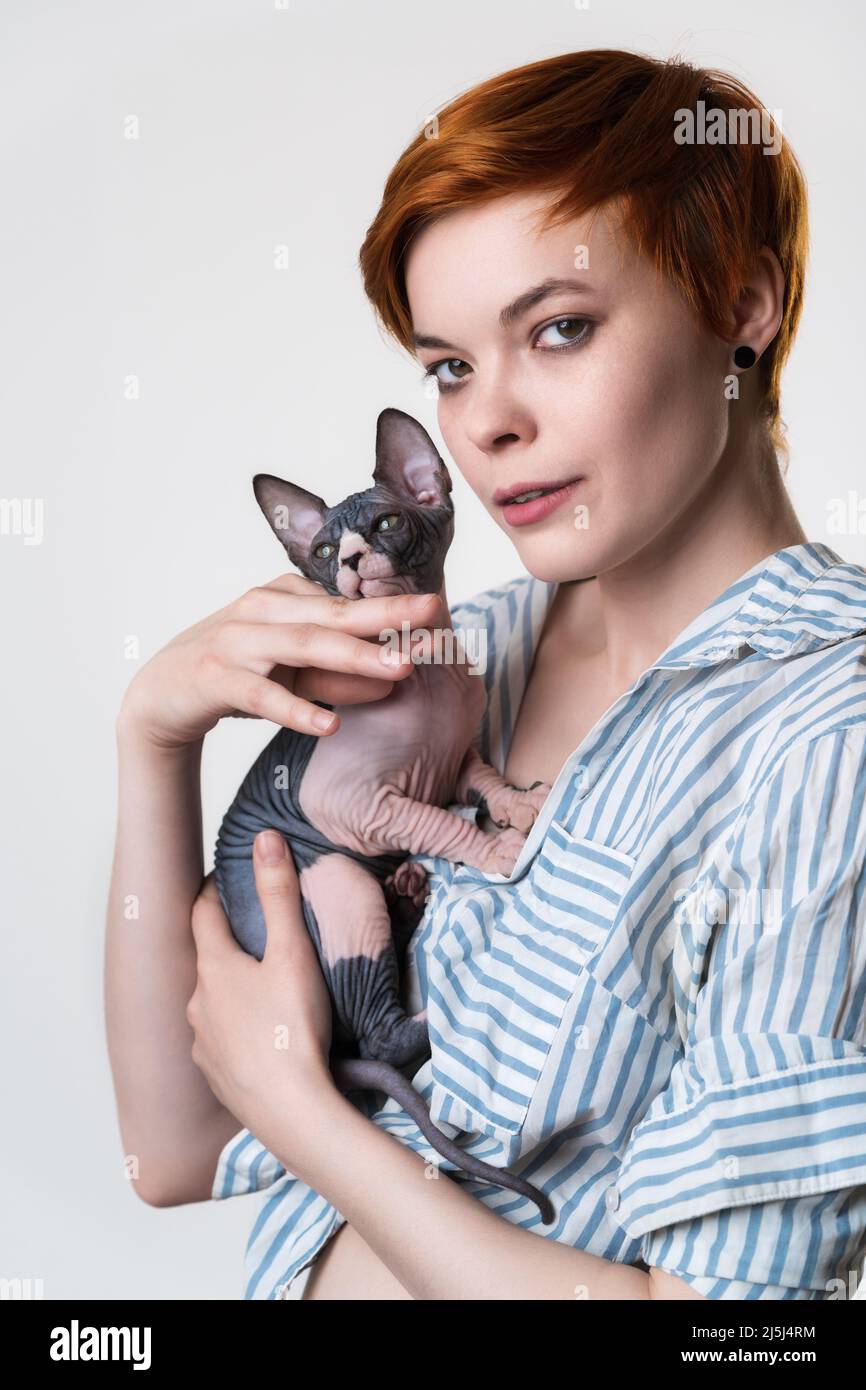 Porträt einer jungen Rotschopf-Frau, die Sphynx Cat in den Händen hält und die Kamera anschaut. Hipster-Hündin mit kurzen Haaren und gestreiftem weiß-blauem Hemd. Stockfoto