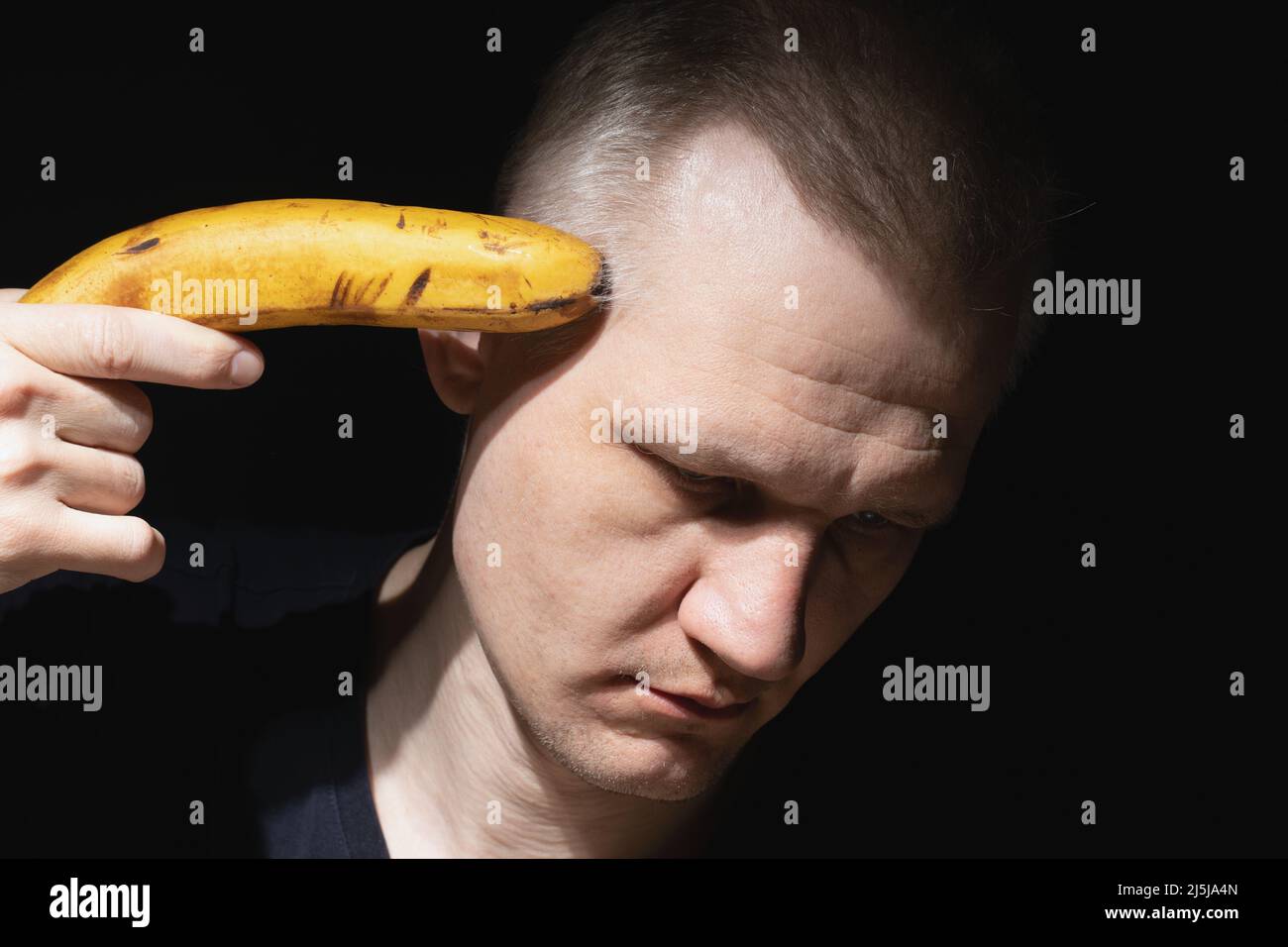 Ein erwachsener Mann mit traurigem Gesicht hält Banane wie eine Pistole auf schwarzem Hintergrund nahe an seinem Kopf. Verzweiflung, Depression und Selbstmordgedanken. Thema psychische Gesundheit. Stockfoto