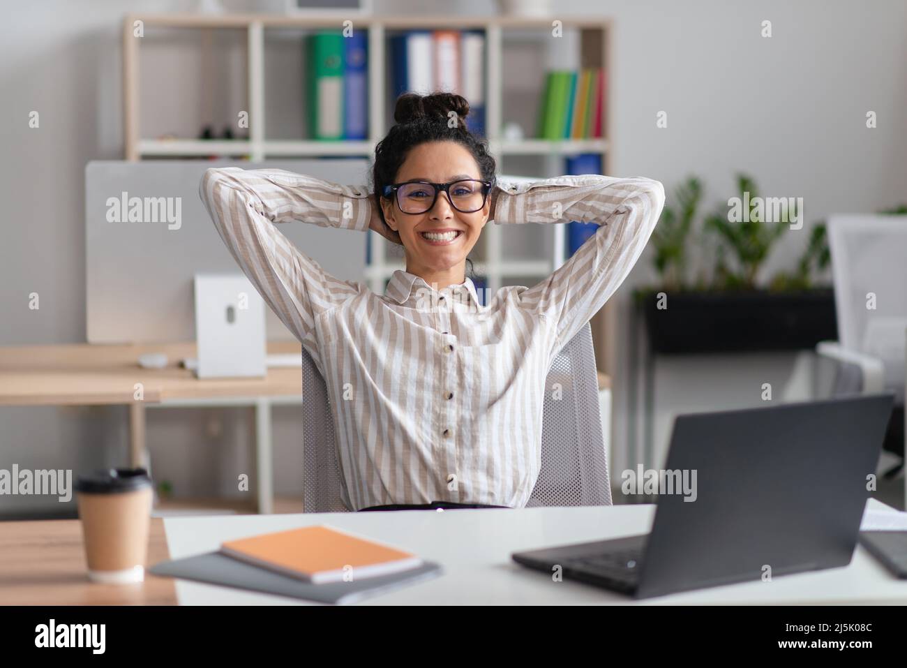 Glückliche junge Angestellte, die sich im Büro vor dem Laptop auf ihrem Stuhl ausruht und sich nach einem erfolgreichen Geschäftsabschluss entspannt Stockfoto