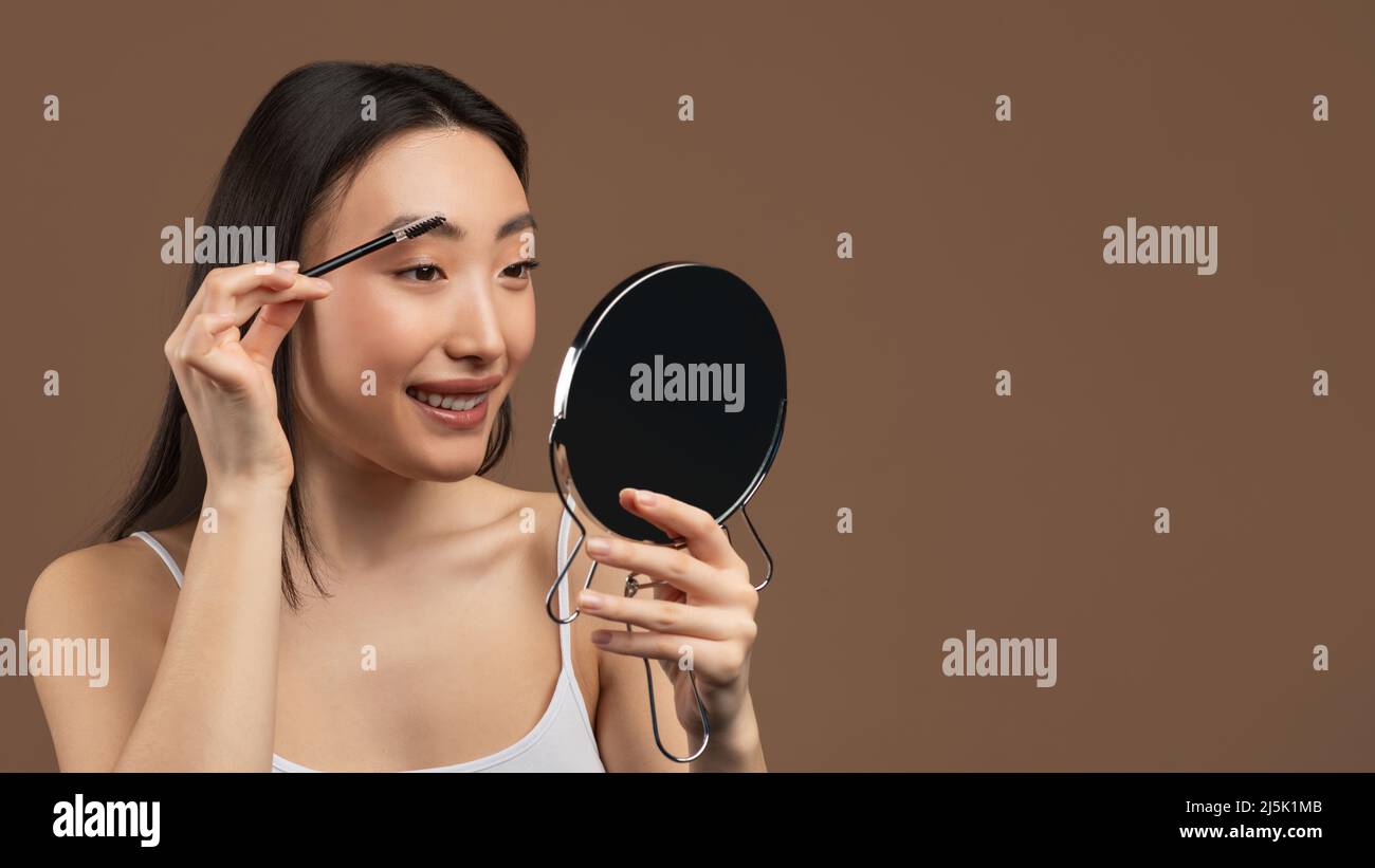 Natürliches Make-up-Konzept. Glückliche asiatische Dame, die Augenbrauen putzt, Pinsel und Spiegel hält, brauner Hintergrund Stockfoto