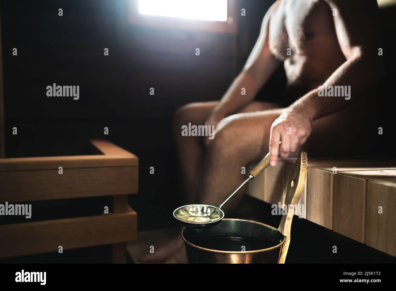 Mann in der Sauna. Finnisches Dampfbad. Person, die Wasser aus dem Eimer auf die heißen Steine gießt. Traditionelle Entspannung und Wellness in Finnland. Stockfoto