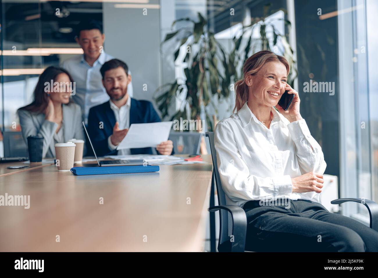 Lächelnde reife Dame sitzt auf einem Stuhl und telefoniert während der Besprechung im modernen Büro Stockfoto