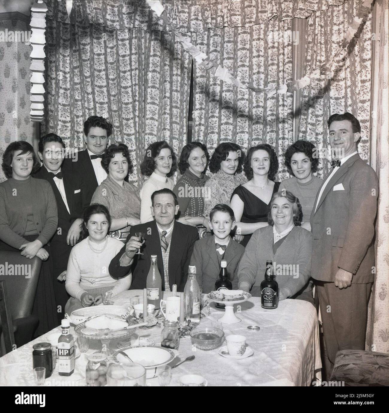 1961, historisch, feiern eine besondere Familienfeier und verkleidet für eine Party, Männer und Frauen unterschiedlichen Alters versammeln sich für ein Gruppenbild am Esstisch, Stockport, Manchester, England, Großbritannien. Auf dem Esstisch sind Marken der damaligen Zeit wie Edmondson's Lemonade und HP Sauce zu sehen. Stockfoto