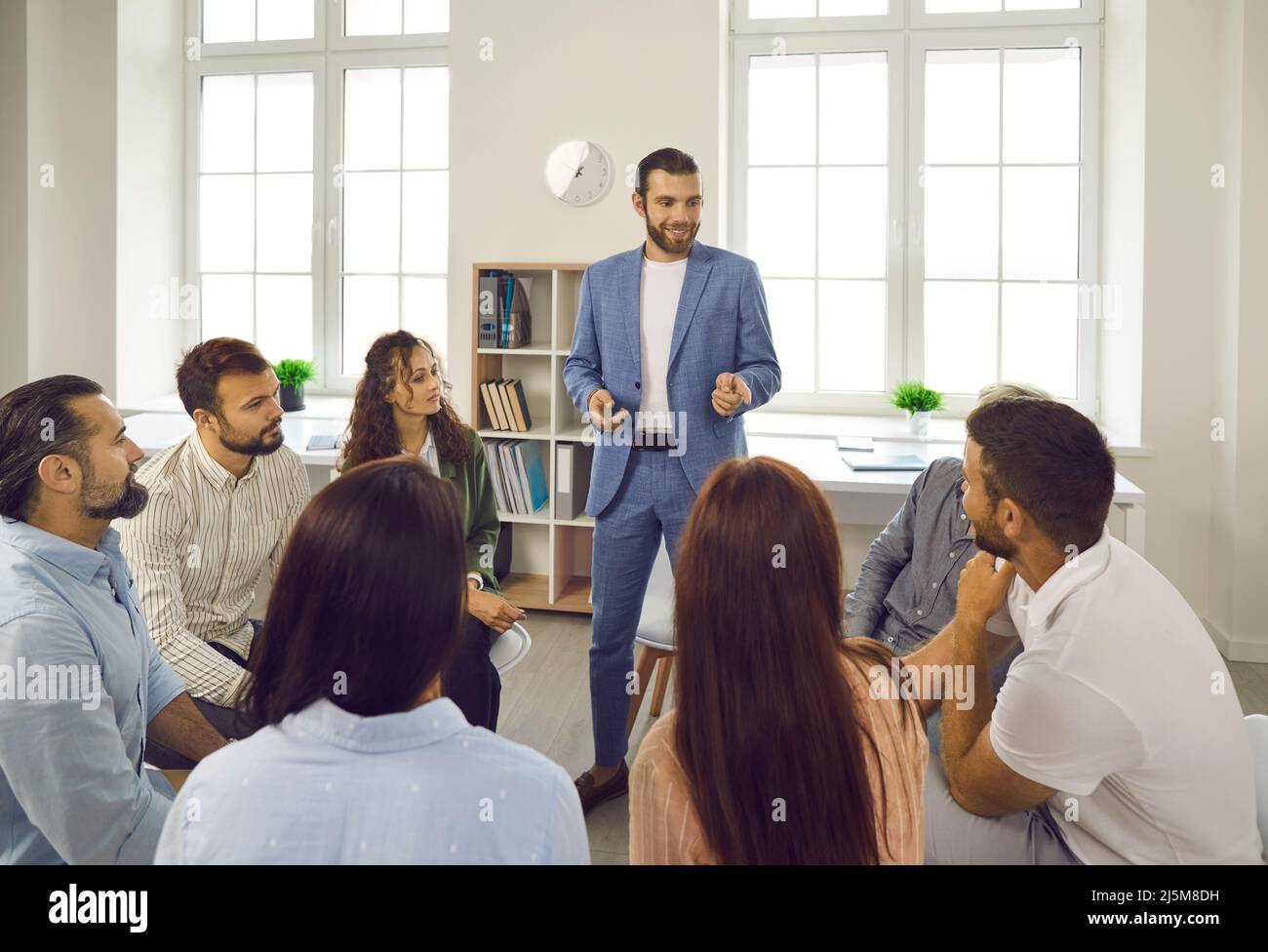 Gruppe von verschiedenen Personen, die im Kreis sitzen und ihrem männlichen Kollegen bei der Besprechung aufmerksam zuhören. Stockfoto