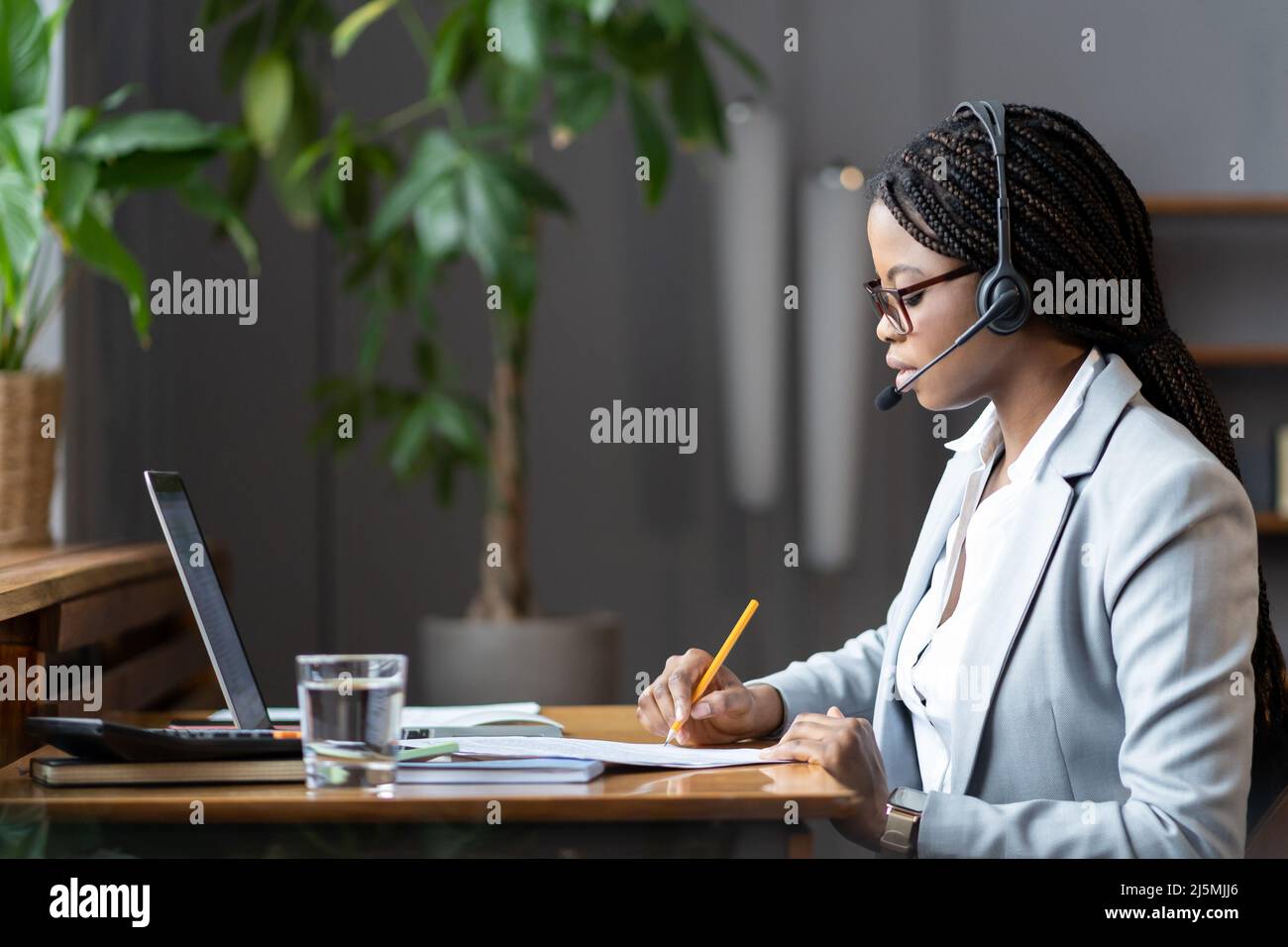 Fokussierte virtuelle Assistentin in afrikanischem Zuhause, die während eines Online-Meetings Notizen macht Stockfoto