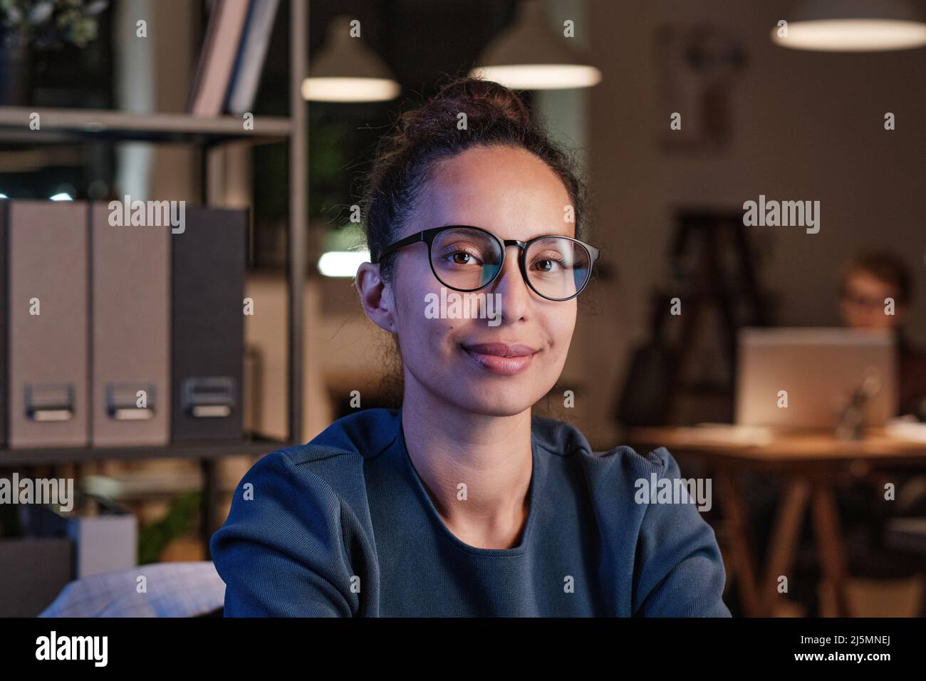 Porträt einer lächelnden attraktiven jungen Geschäftsfrau aus dem biracial Bereich in einer Brille, die im Büro an den Regalen sitzt Stockfoto