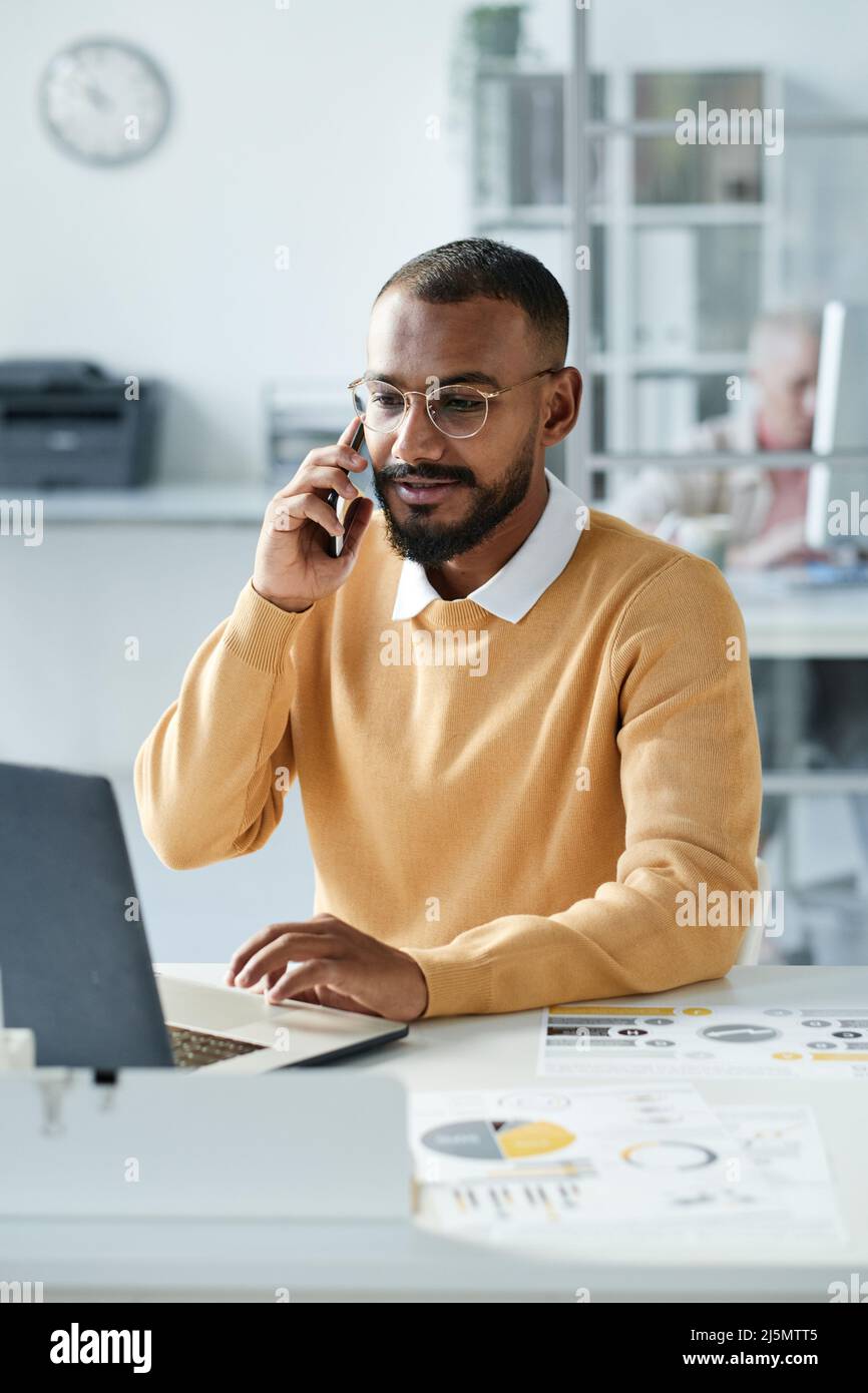 Inhalt selbstbewusster junger multiethnischer Analytiker für Brillen, der mit Laptop und Dokumenten am Schreibtisch sitzt und telefoniert Stockfoto