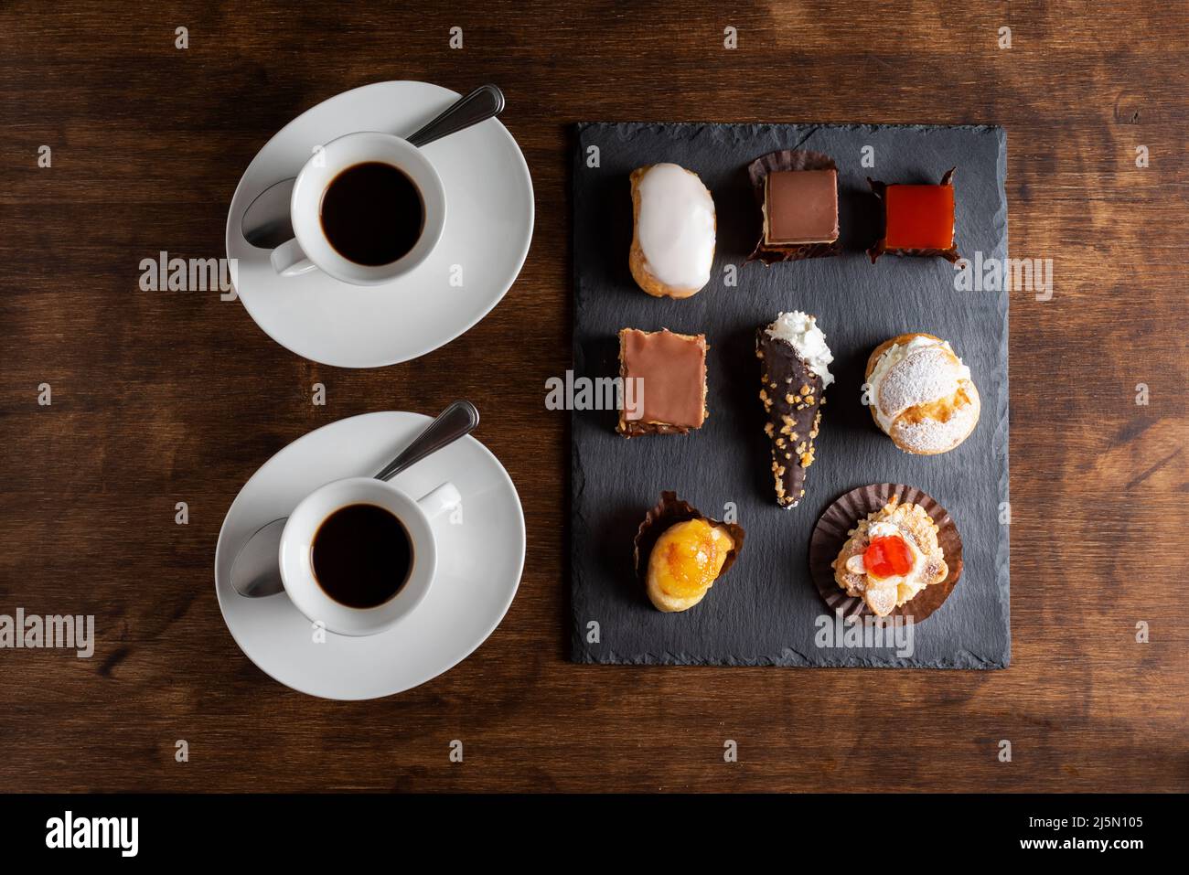 Kleine Kuchen auf einem rustikalen Steinteller, neben einigen Kaffees auf einem alten Holztisch. Stockfoto