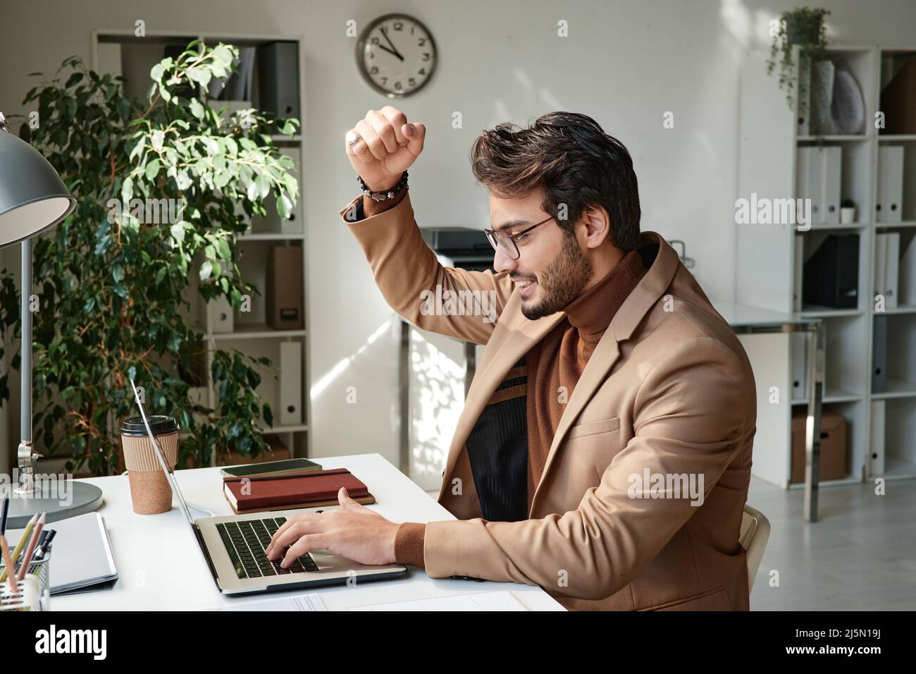 Lächelnder, gutaussehender Manager aus dem Nahen Osten in Brillen, der mit Ja-Gesten und einem Laptop unterwegs war, während er ein Ergebnis erzielt hat Stockfoto