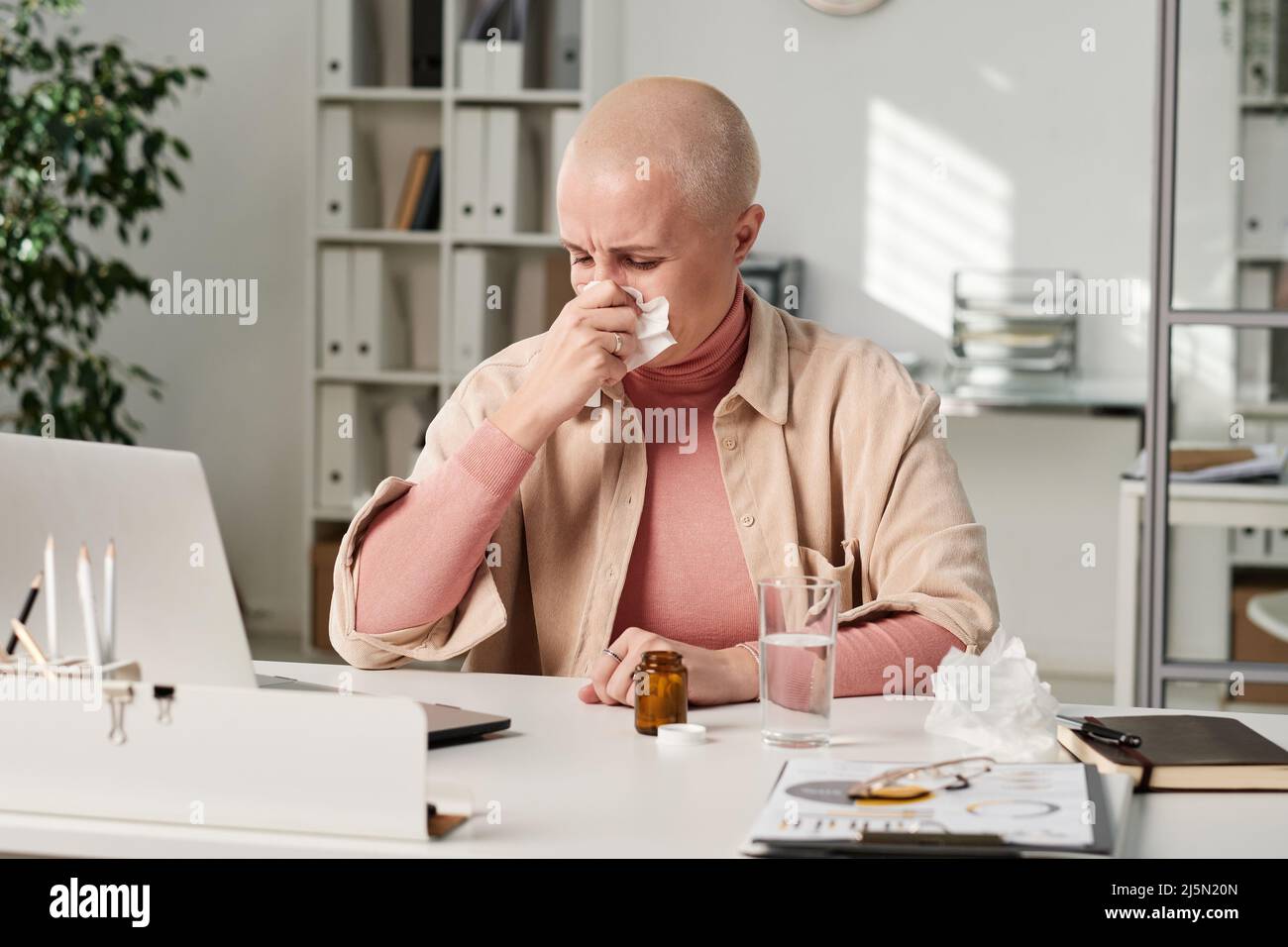 Junge kahle Geschäftsfrau wischte sich mit Serviette die Nase, während sie am Arbeitsplatz niesen Stockfoto