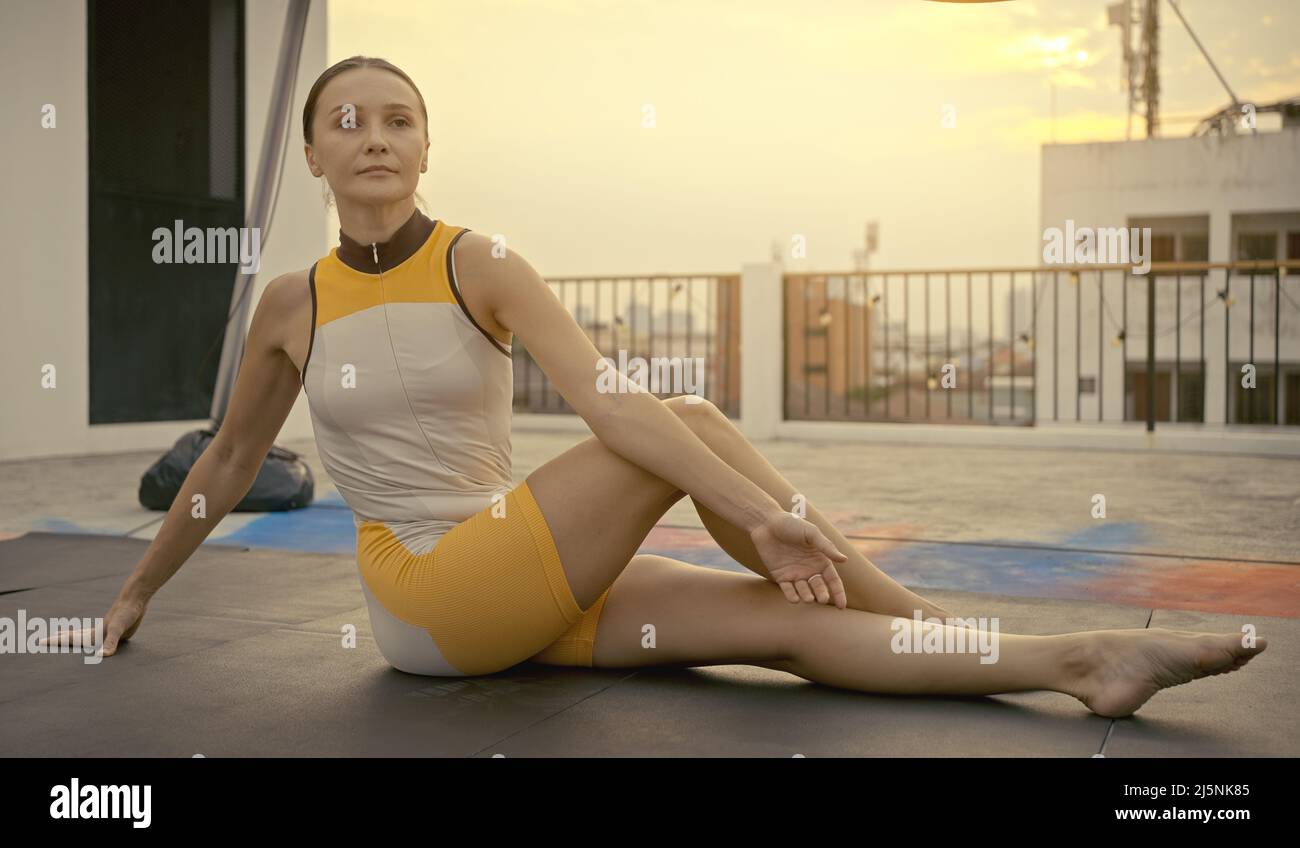 Frau in Sportkleidung, die sich ausdehnt und sich aufwärmt, bevor sie am Abend, wenn die Sonne untergeht, auf dem Dach Yoga macht. Selbstversorgung und Gesundheitskonzepte. Stockfoto