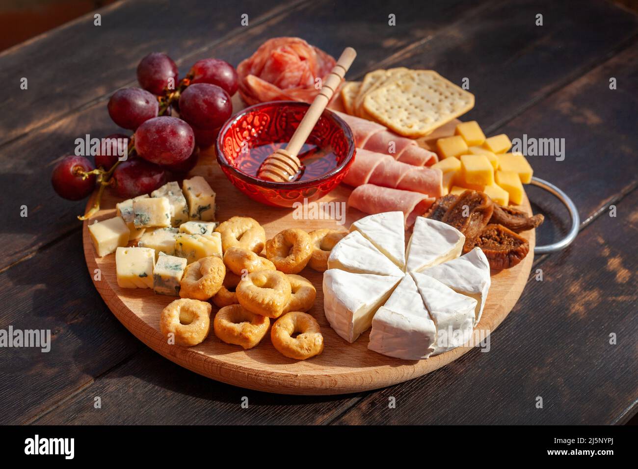 Rundes Schneidebrett mit verschiedenen Käsesorten, Wurstrosette, Schinken, Cracker, Trauben und Feigen mit Honig. Aufschnitt und Käseplatte auf dem Tter Stockfoto