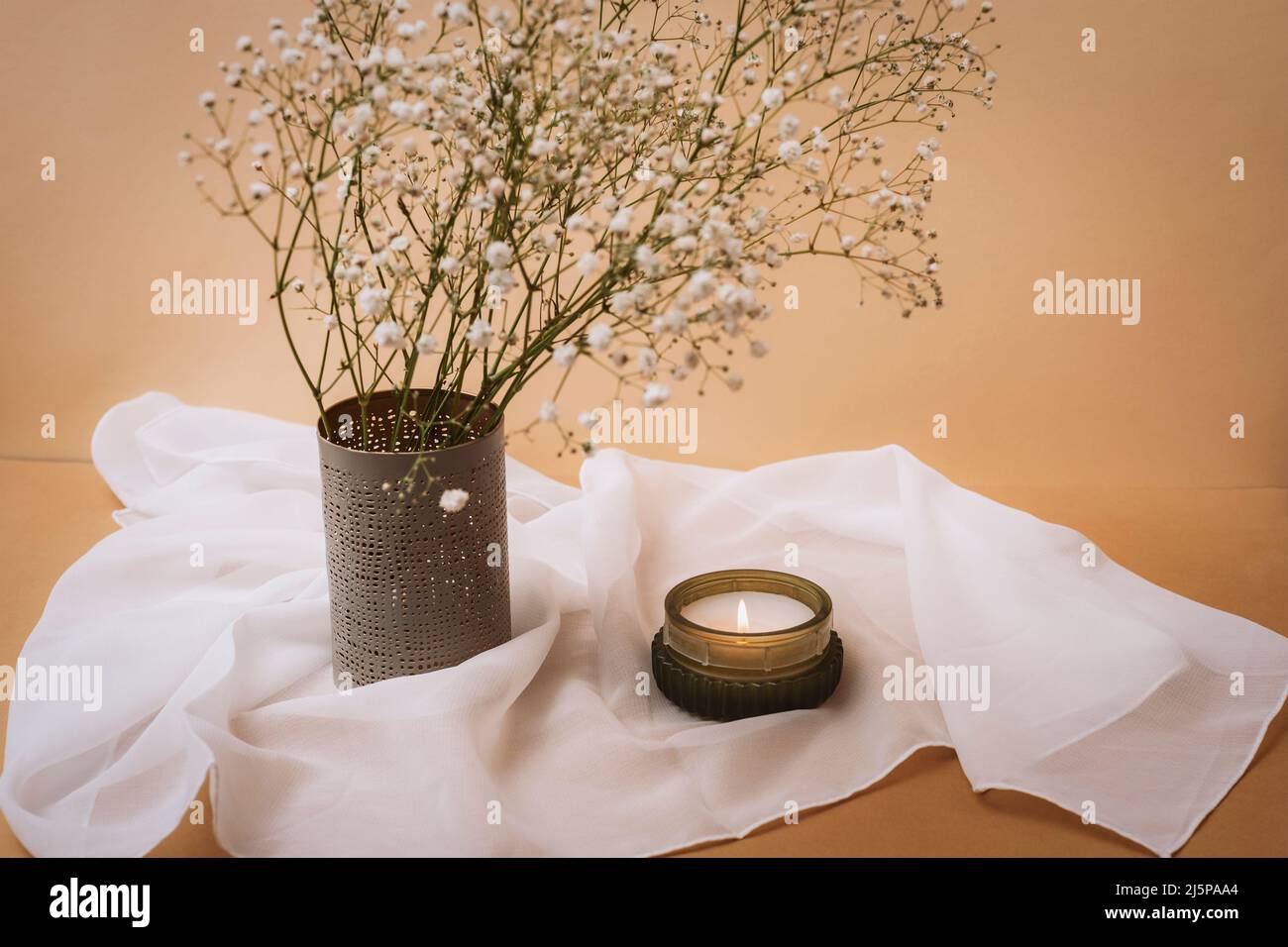 Kerze und Gypsophila Blumen in Vase auf weißer Tischdecke auf beigem Tisch. Stillleben. Stockfoto