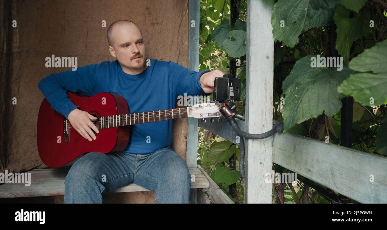 Mann mit Gitarre sitzt auf der Veranda und stimmt das Instrument mit einer App am Telefon ab Stockfoto